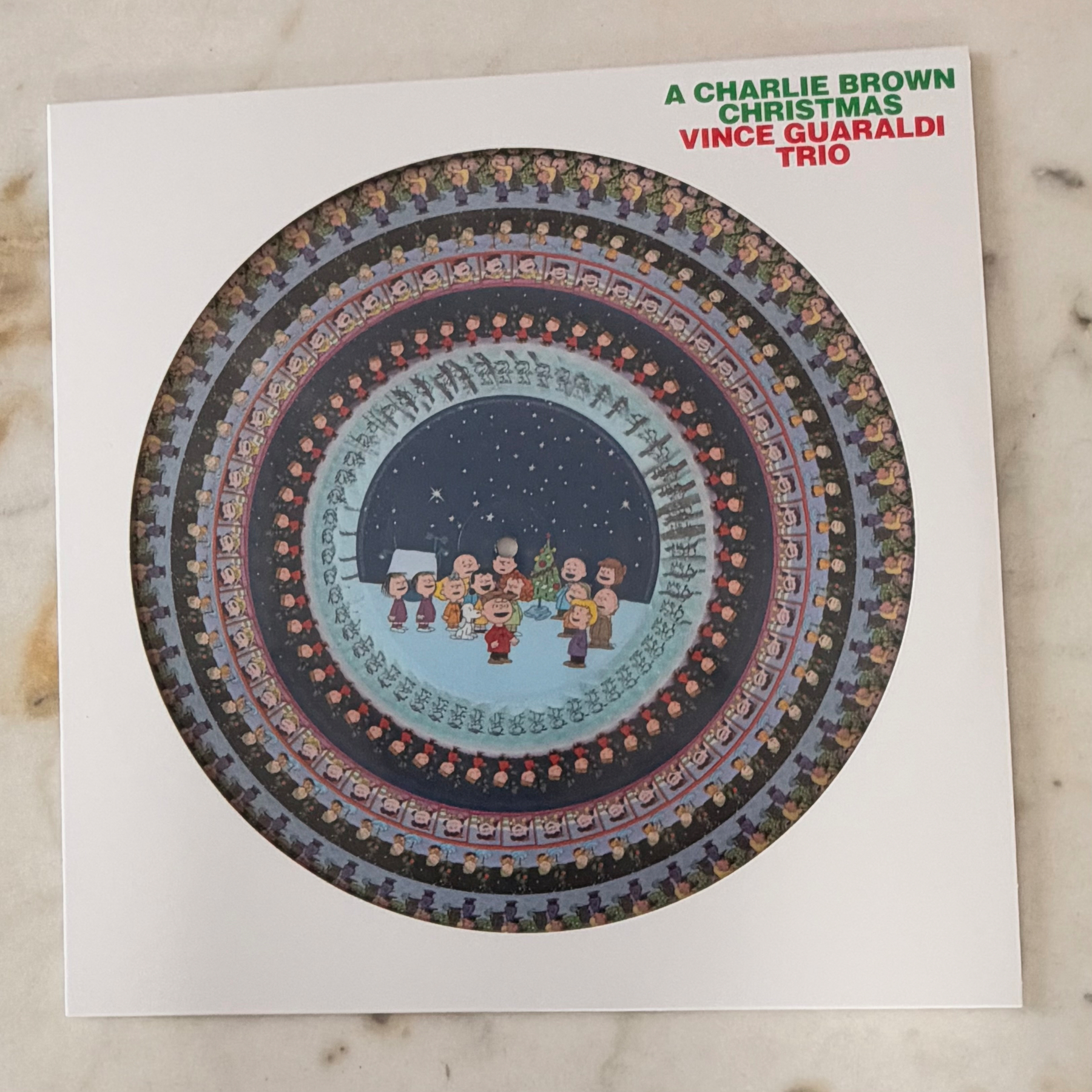 Auto-generated description: A Christmas-themed album cover featuring the Vince Guaraldi Trio with circular, repeating images of Peanuts characters singing around a snowy scene.