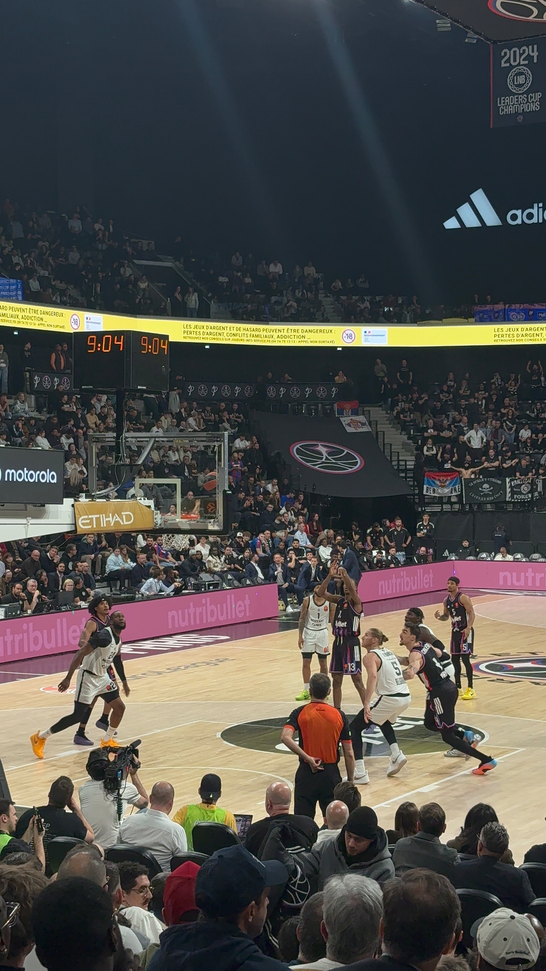 A basketball game is in progress with players actively competing on the court surrounded by a large audience.