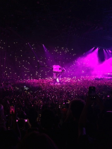 A large crowd is attending a concert with many people holding up their phones, while purple stage lights illuminate the performer.