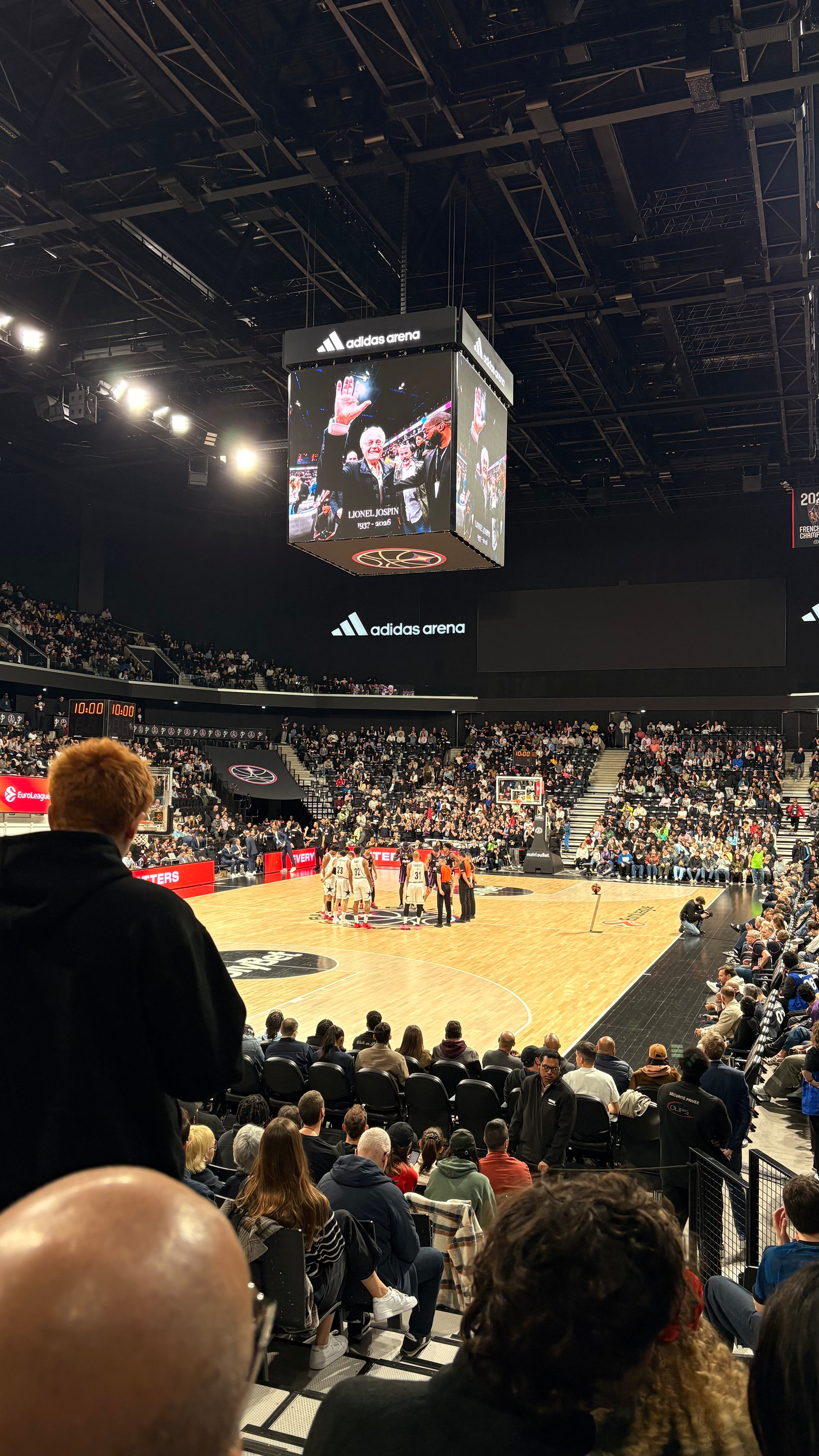 A basketball game is taking place in an indoor arena filled with spectators, featuring a large scoreboard hanging above the court.