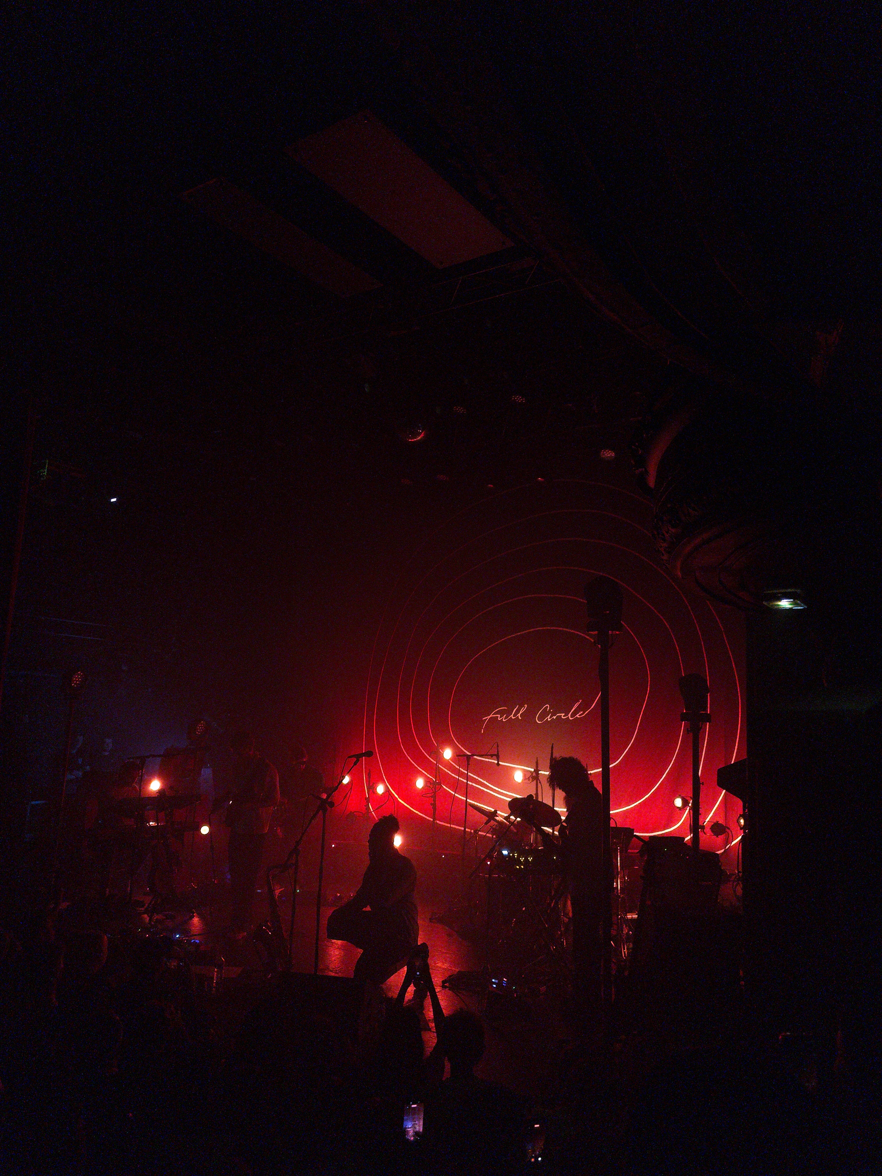 Auto-generated description: A band performs on a dimly lit stage with red lighting and circular patterns in the background.