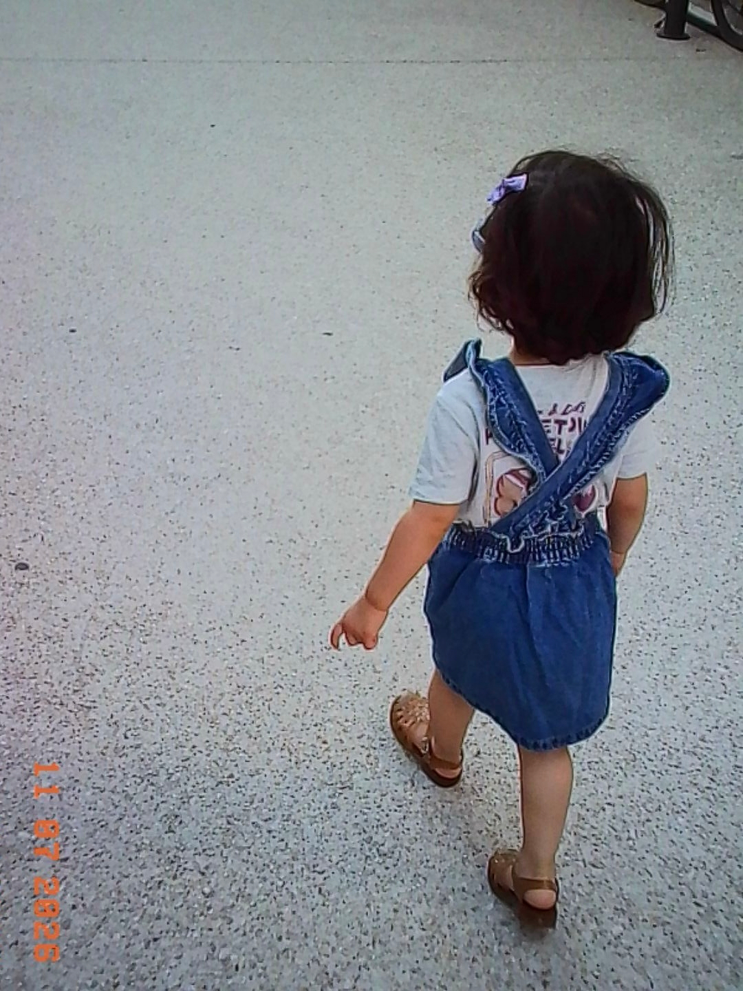 A young child with a hair clip is walking on a speckled floor wearing a blue denim overall dress and brown sandals.