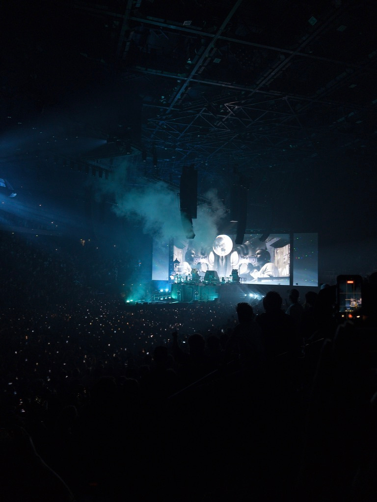 A large crowd watches a live concert in a dark venue with bright stage lights, fog effects, and a giant screen displaying black-and-white visuals.