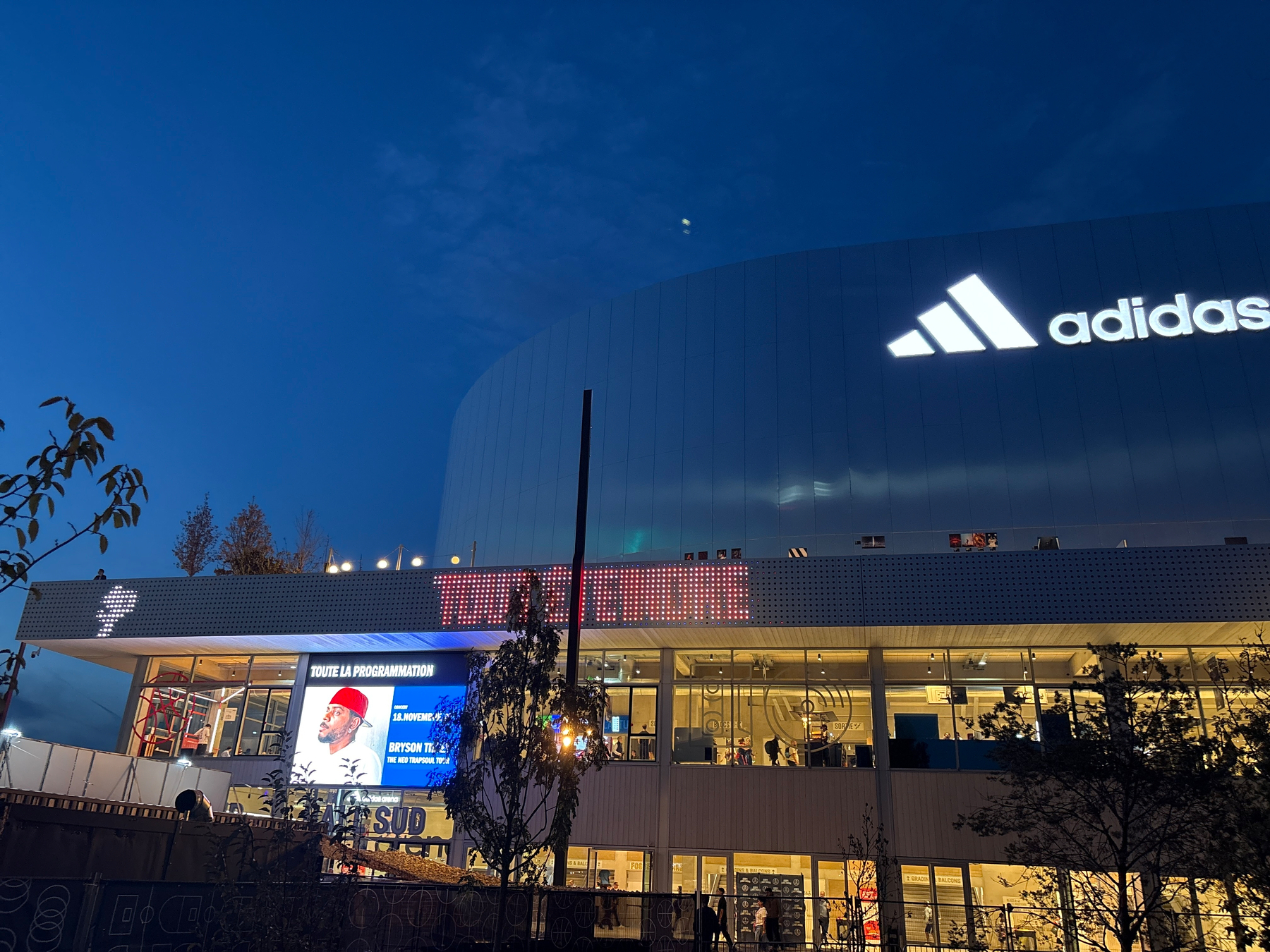 A large Adidas-branded building with illuminated signage and a digital screen displaying an event or program information is shown at dusk.