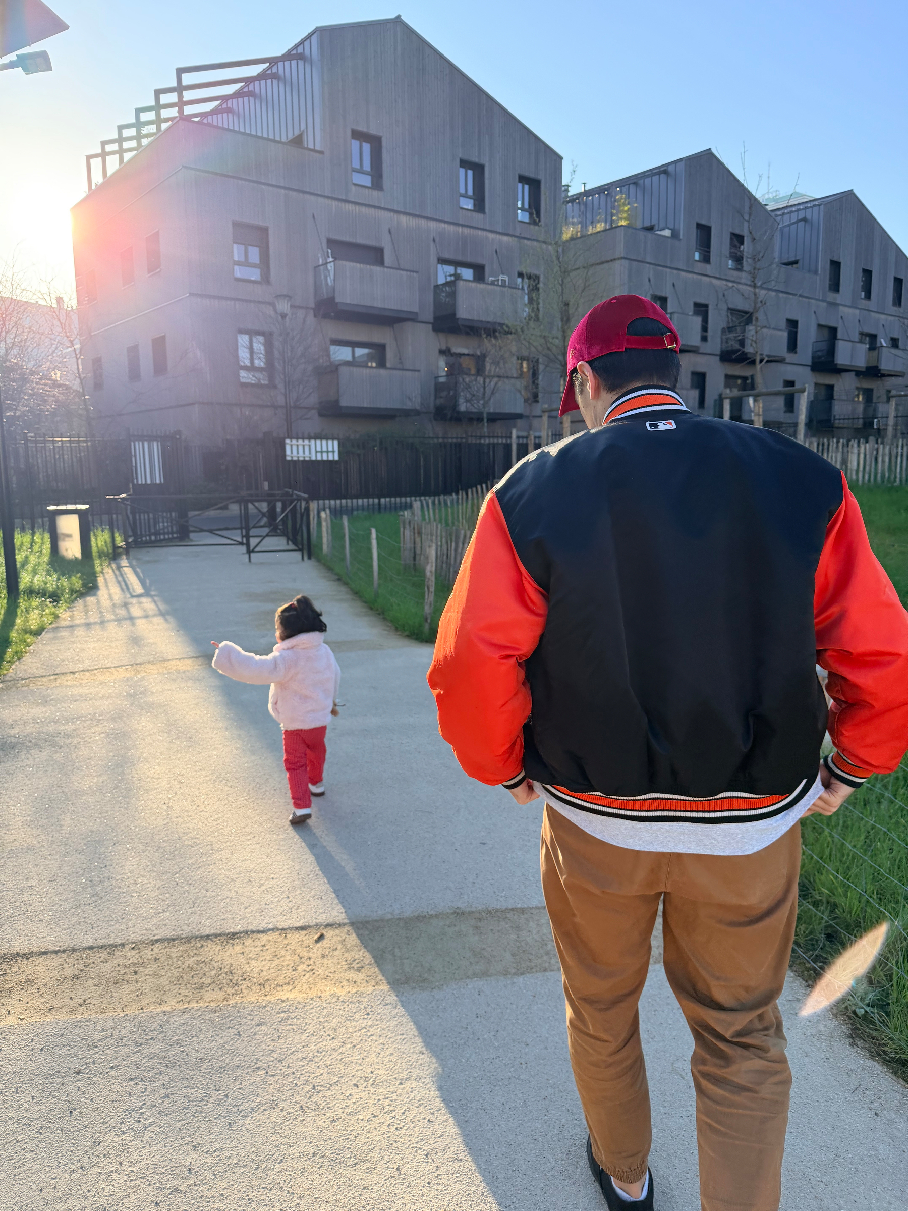 A person wearing an orange and black jacket walks with a child down a sunlit path in a residential area.