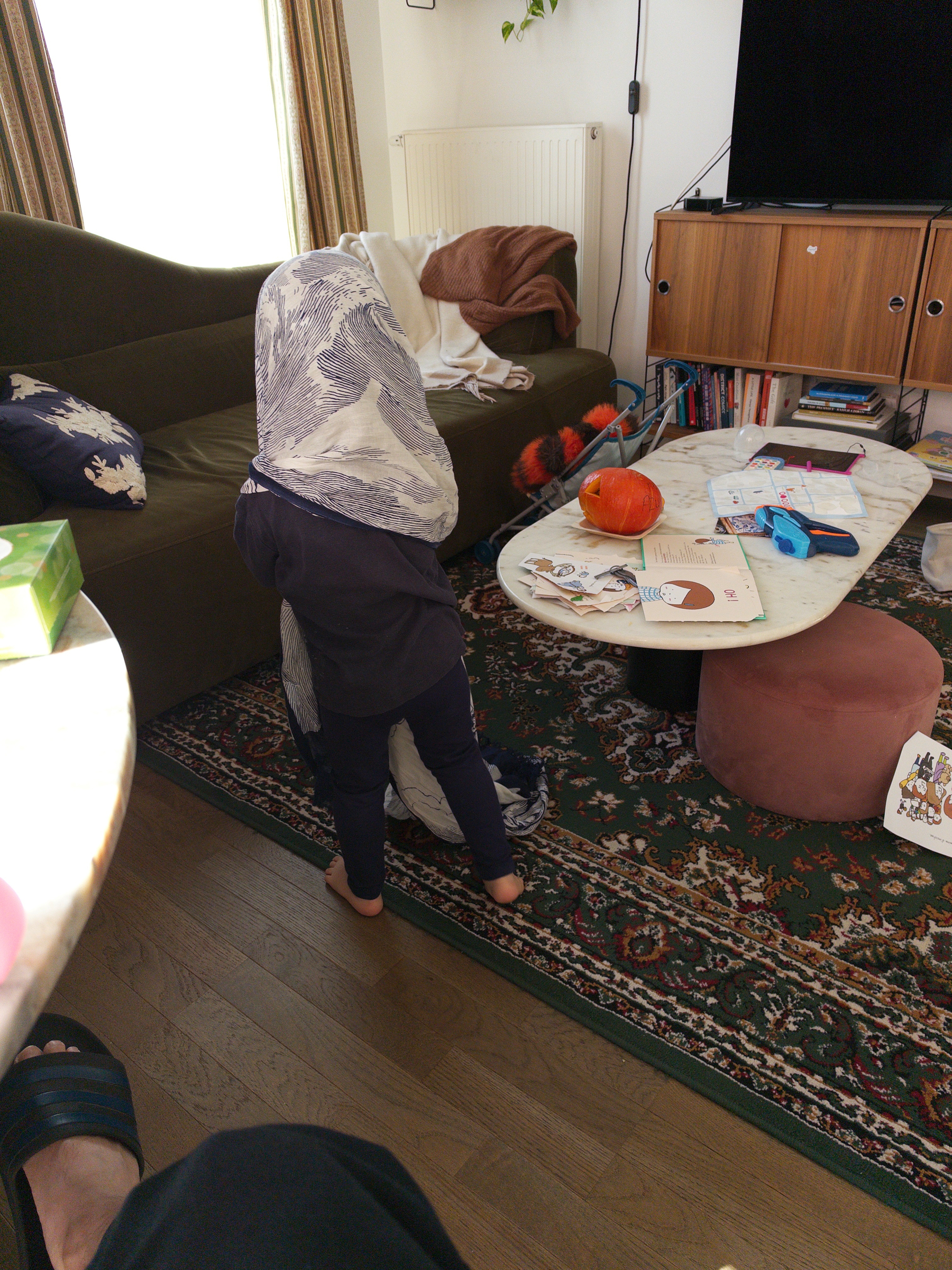 Auto-generated description: A child is standing in a living room with a piece of white and blue cloth draped over their head, surrounded by furniture, books, and various items on a coffee table.