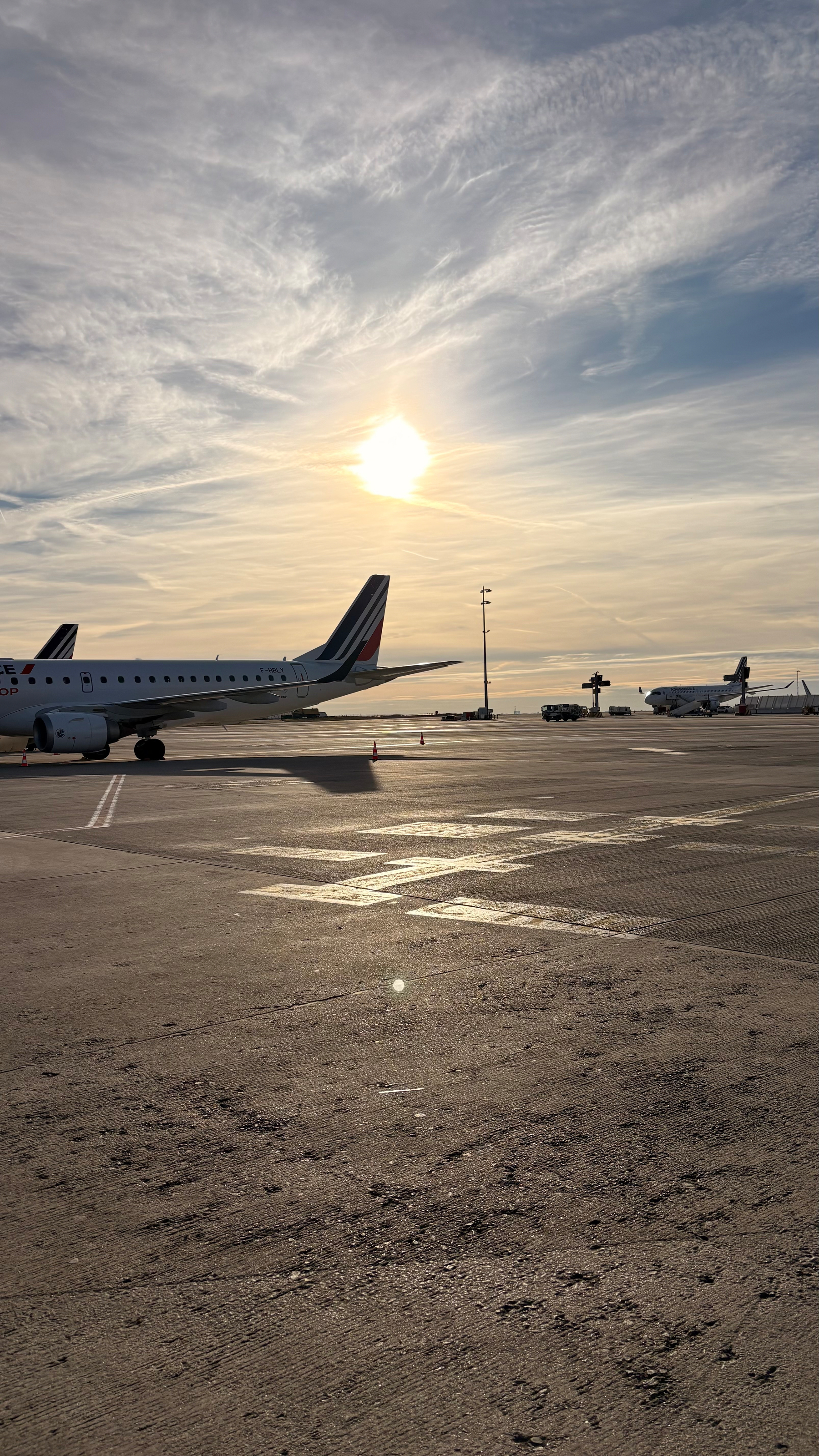 Airplanes are parked on an airport tarmac under a cloudy sky with the sun setting or rising.