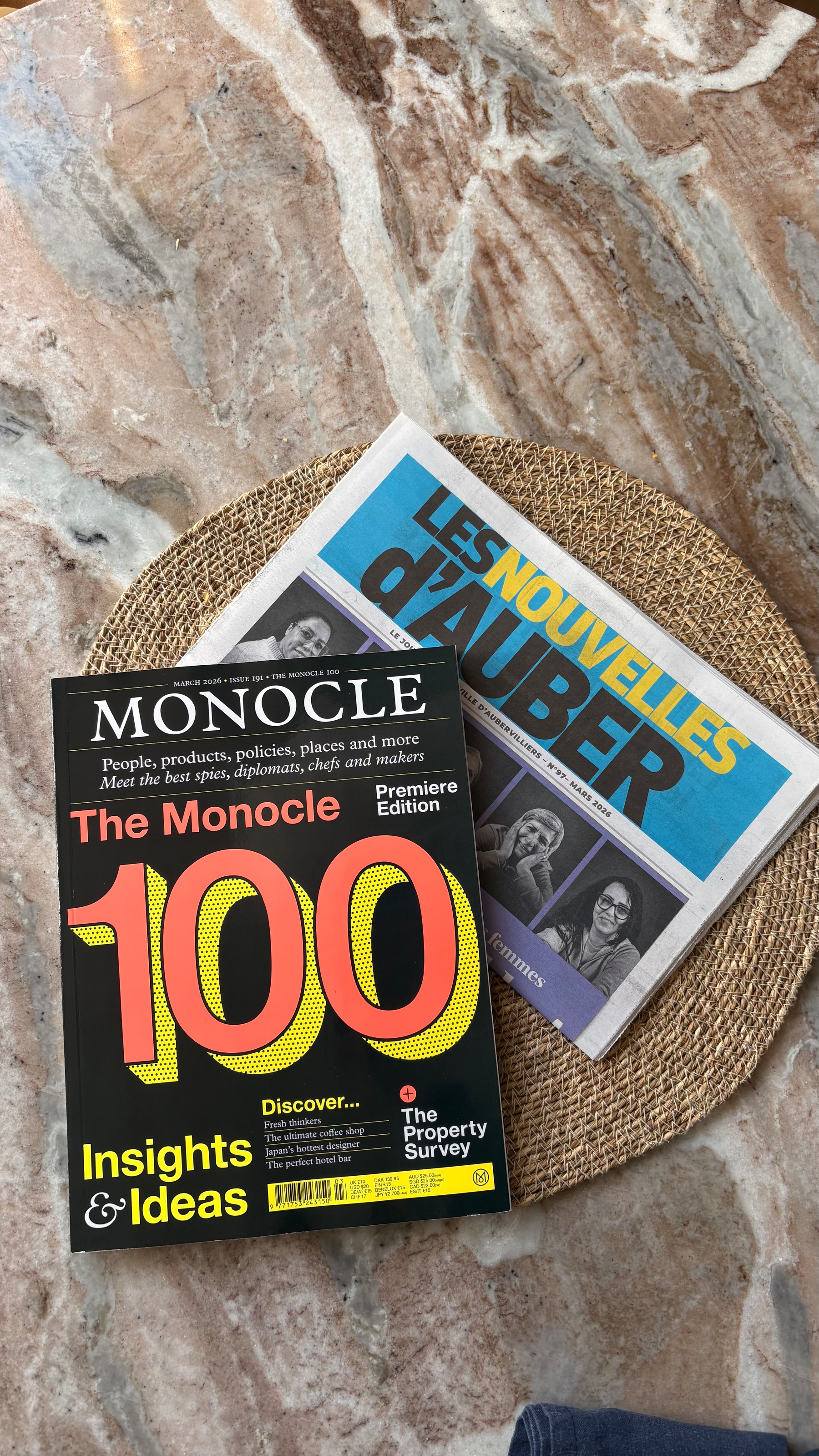 A coffee table holds a copy of Monocle magazine and a newspaper titled Les Nouvelles d'Auber on a woven mat.