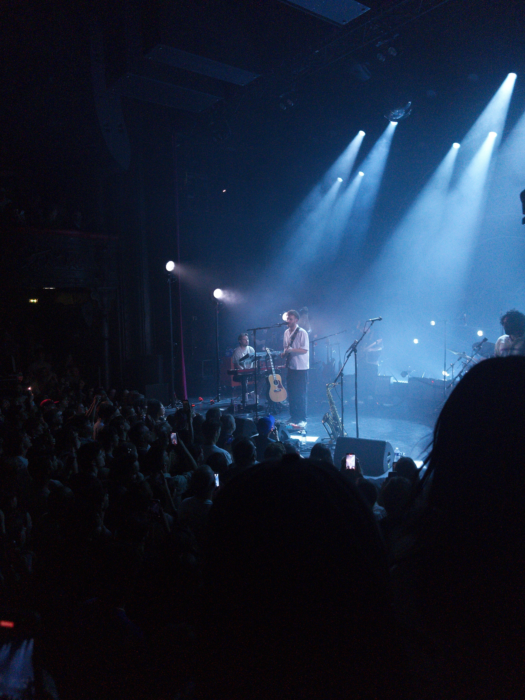 Auto-generated description: A musician performs on stage in front of a large crowd with blue stage lighting and instruments around.