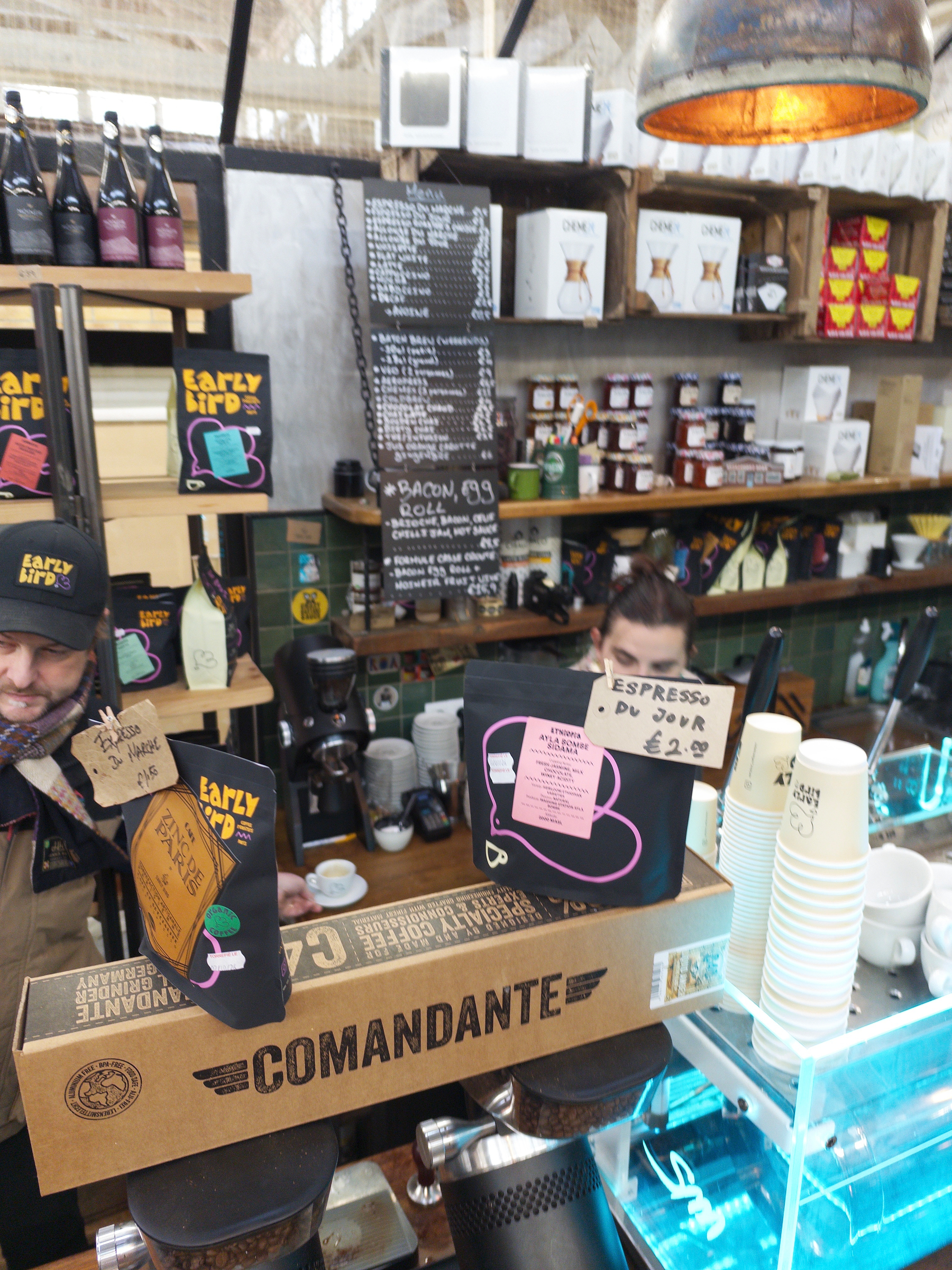 A coffee shop counter displays various coffee bags, cups, and a handwritten sign with prices, while a barista works behind the counter.