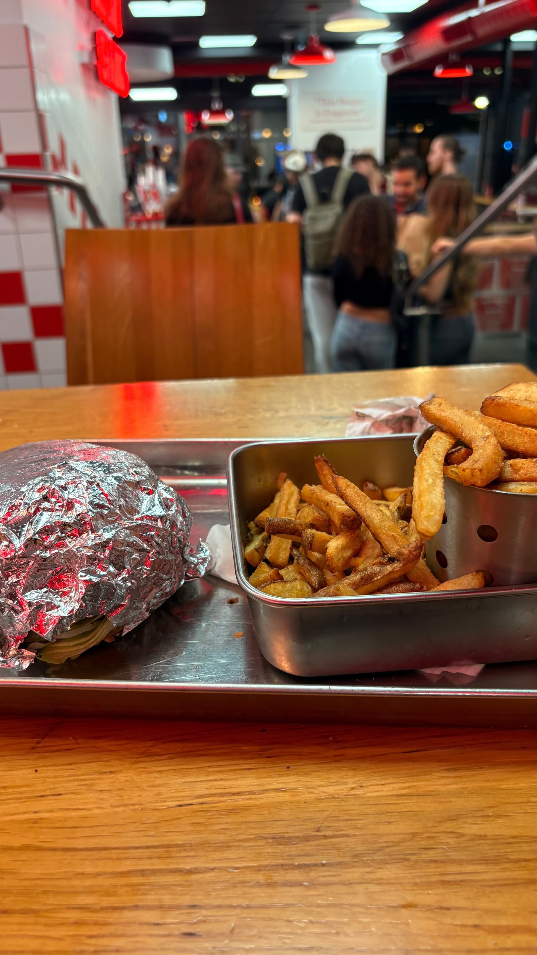 Auto-generated description: A wrapped burger and a metal tray filled with fries and curly fries sit on a wooden table in a busy restaurant.