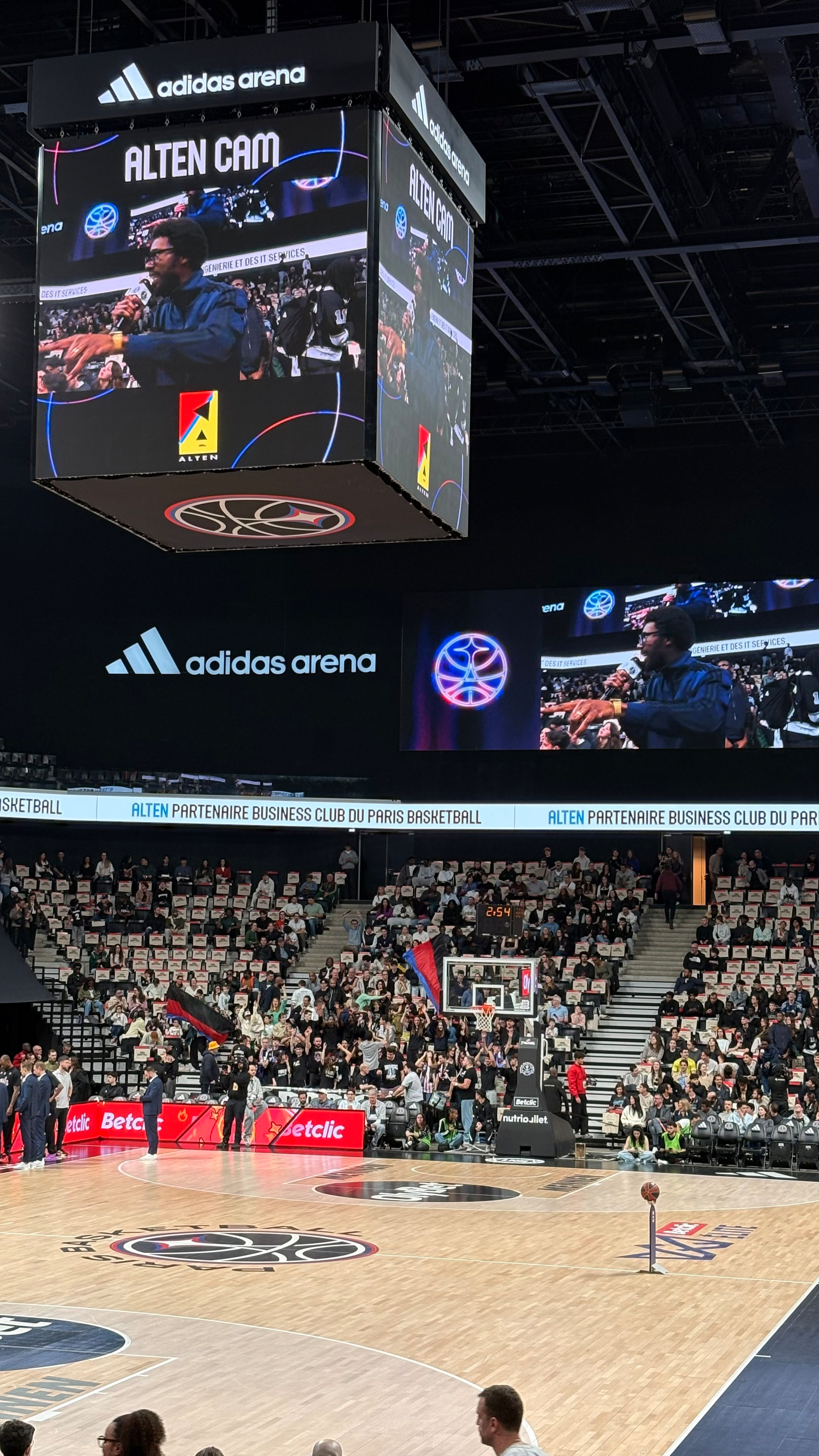 Inside the adidas arena, a basketball game is viewed from the stands, with a large screen displaying the event and spectators in the background.