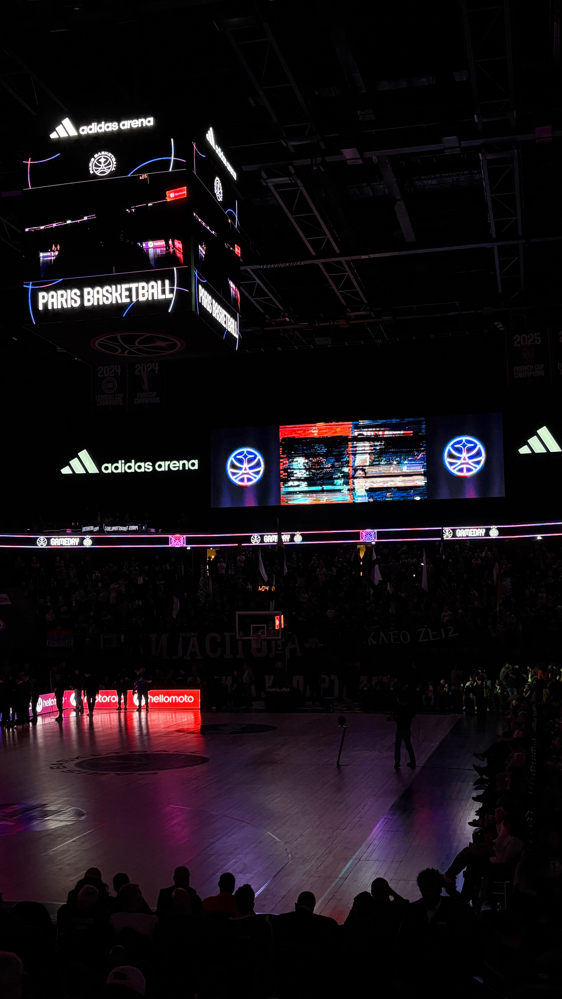 A dimly lit basketball arena features a central scoreboard displaying PARIS BASKETBALL with spectators surrounding the court.
