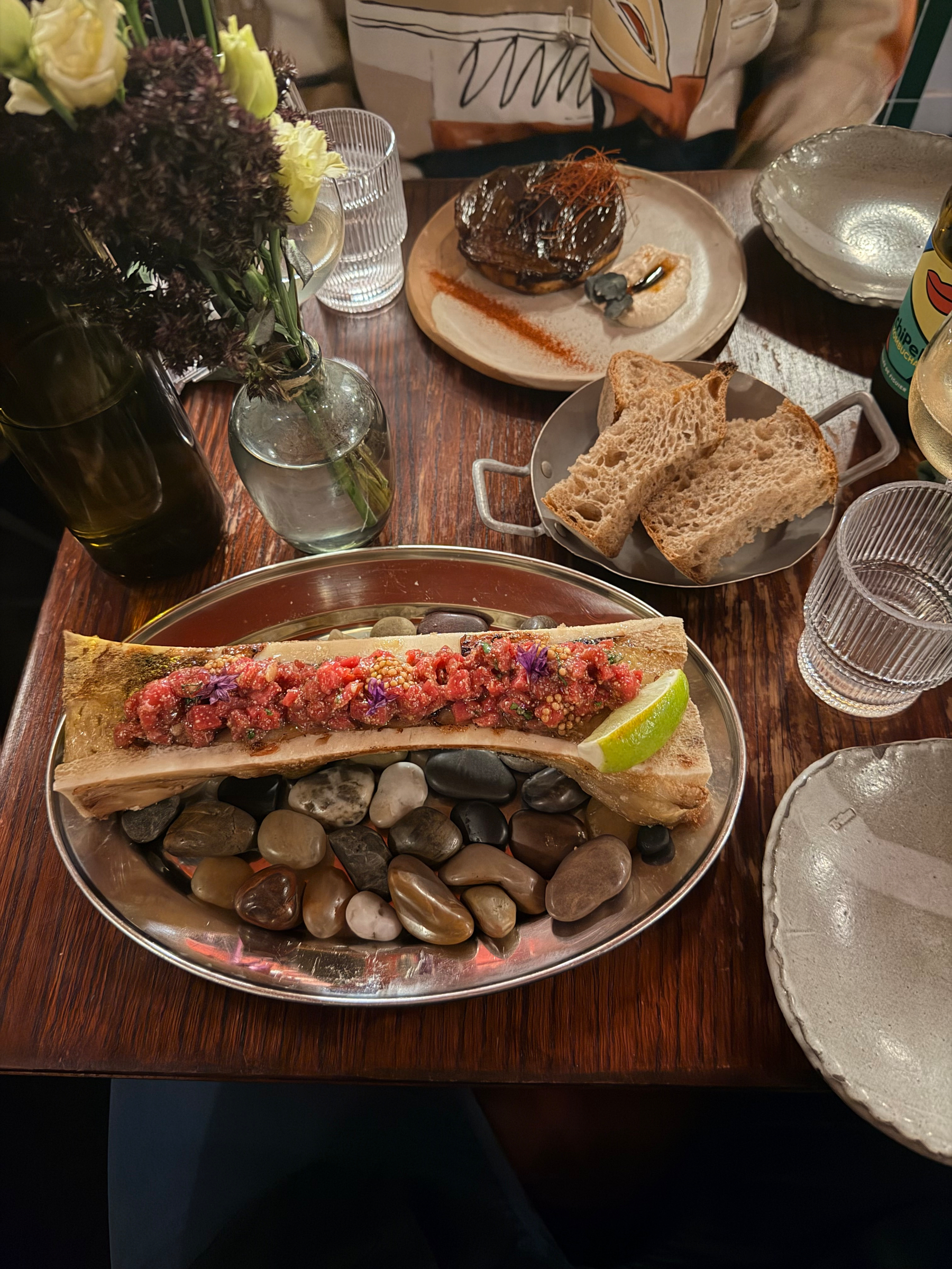 A beautifully plated dish featuring a bone marrow with colorful toppings is accompanied by sliced bread and surrounded by decorative stones on a wooden table setup.
