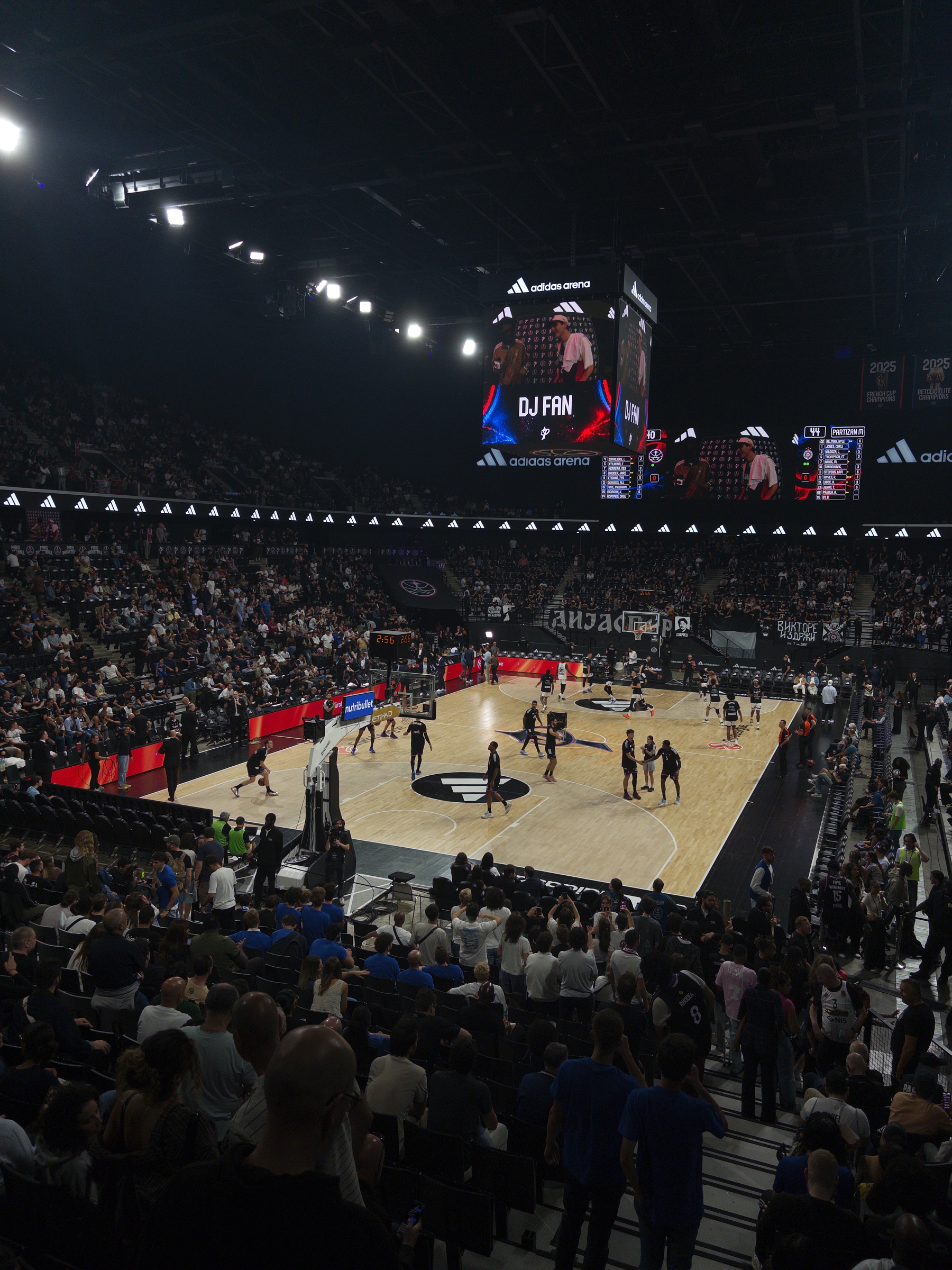 A basketball game is taking place in a large, crowded indoor arena with players on the court warming up or playing and spectators filling the stands.