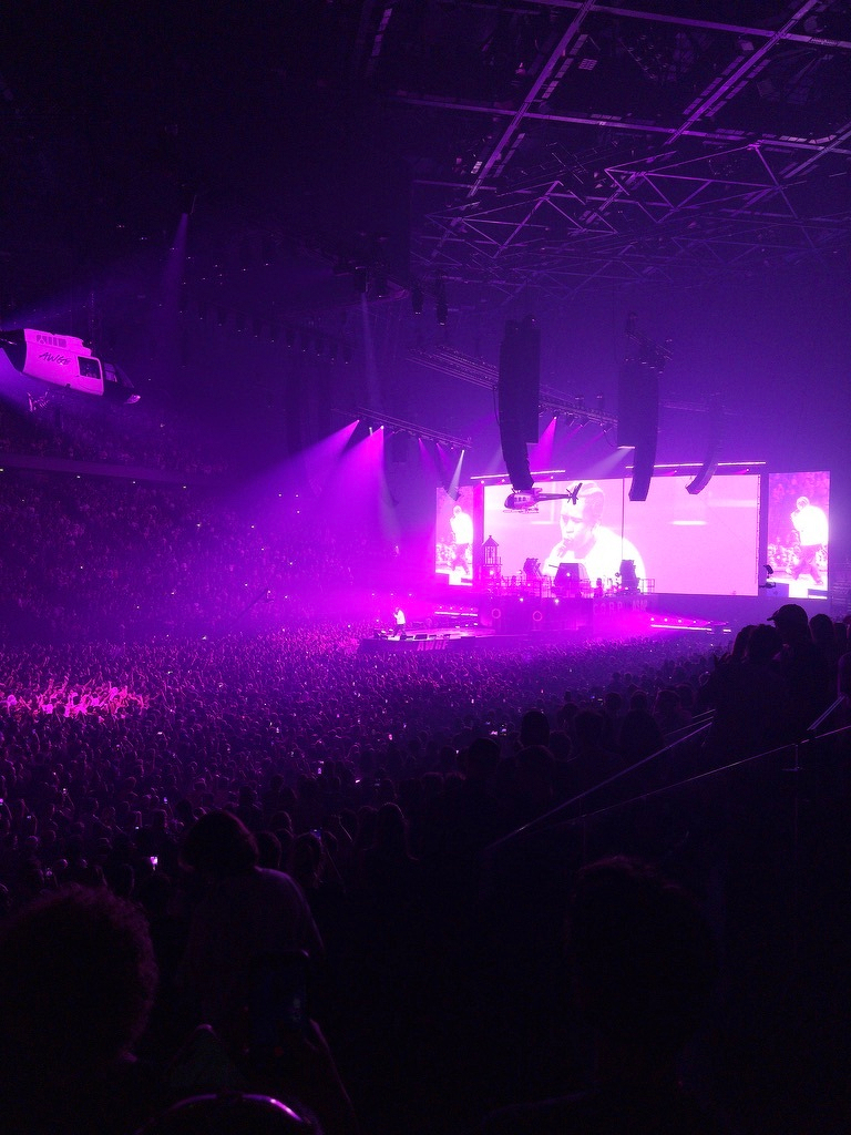 A large crowd enjoys a concert with vibrant purple lighting and a helicopter suspended above the stage.