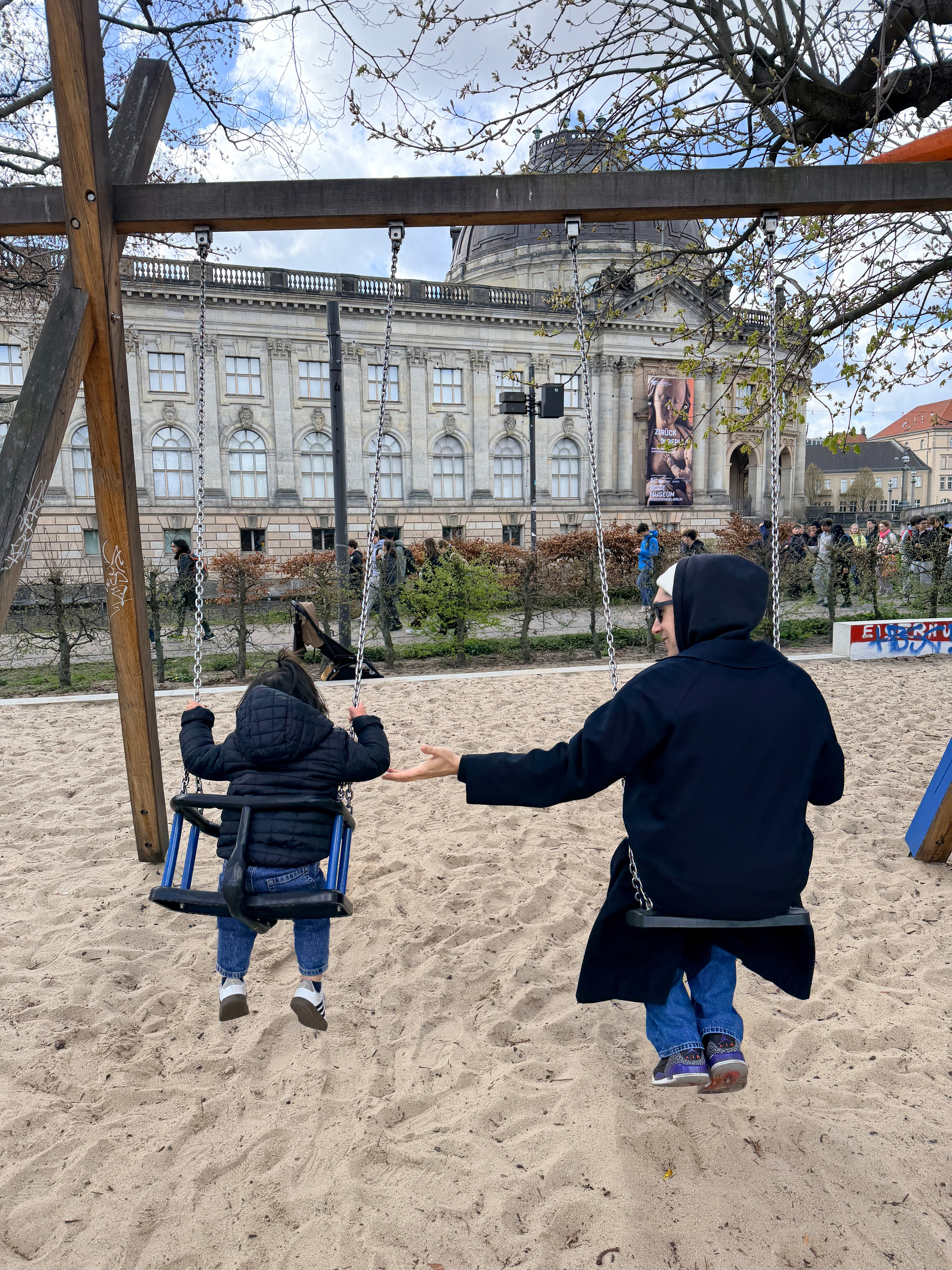 Auto-generated description: A person and a child are on swings at a sandy playground with a large building in the background.