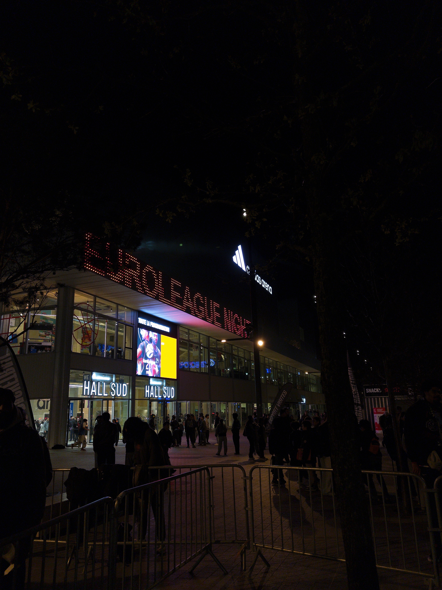 A nighttime scene shows a building with bright signage that reads EUROLEAGUE NIGHT, surrounded by people and illuminated by various lights.