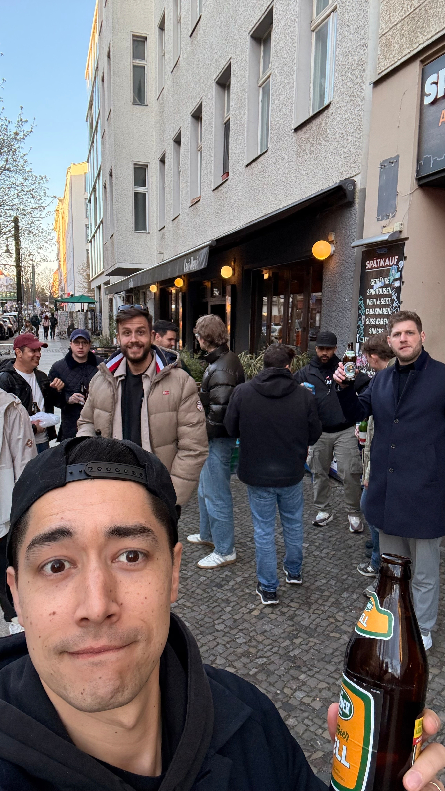 Auto-generated description: A group of people are gathered outside a building on a busy street, with one person holding a bottle in the foreground.