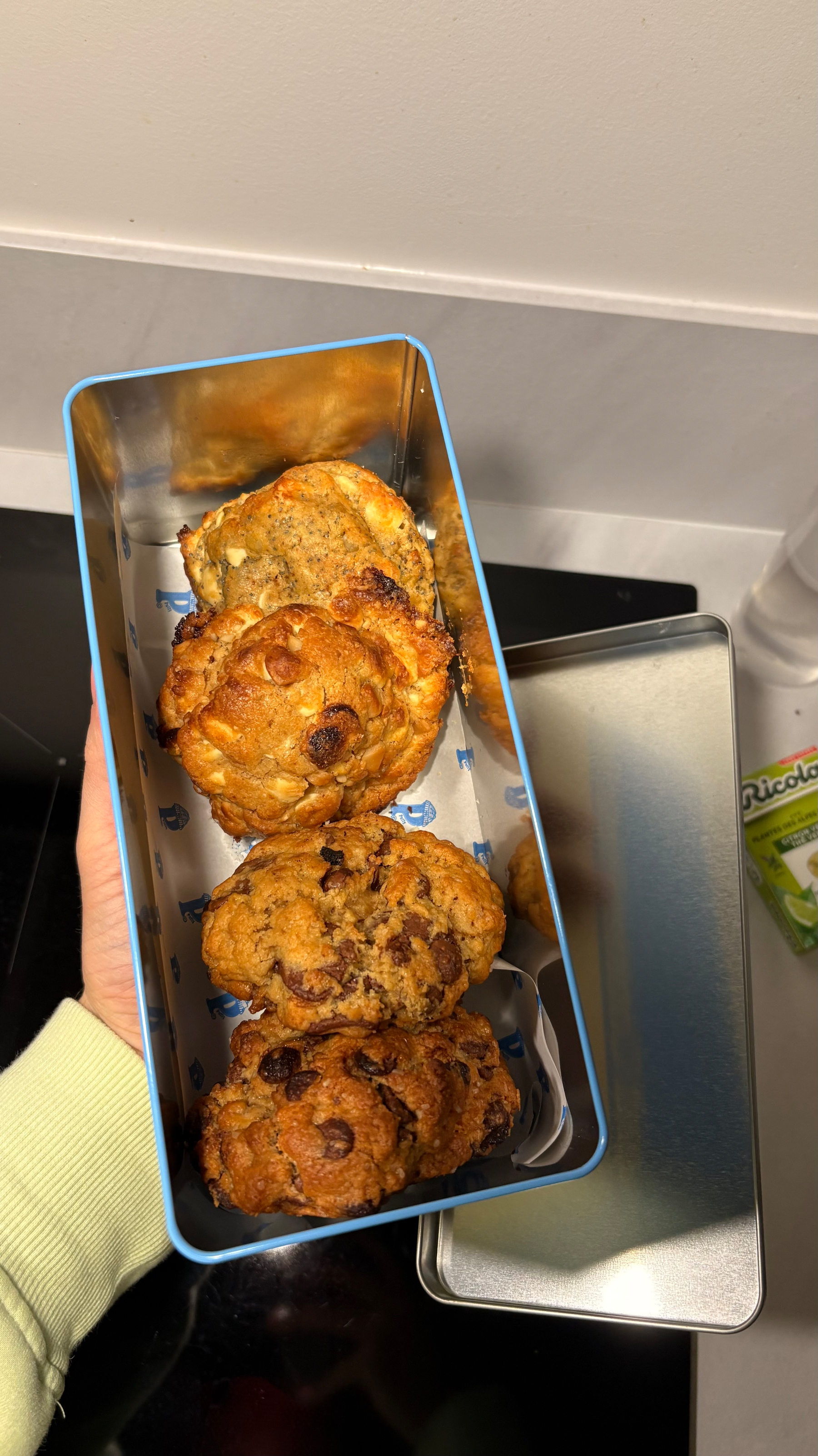 A hand holds an open rectangular tin containing several chocolate chip cookies.