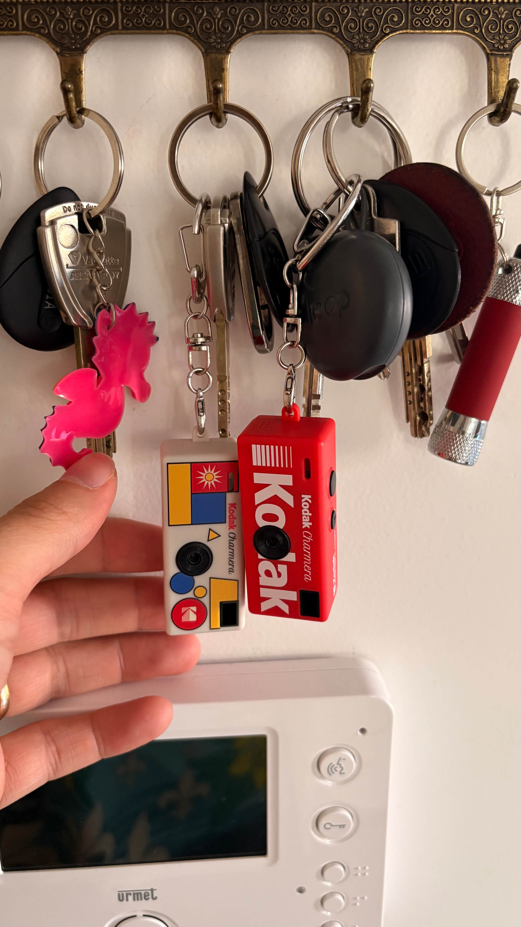 A hand is holding colorful toy keychains attached to metal key rings hanging from an ornate wall-mounted key holder above a white intercom unit.