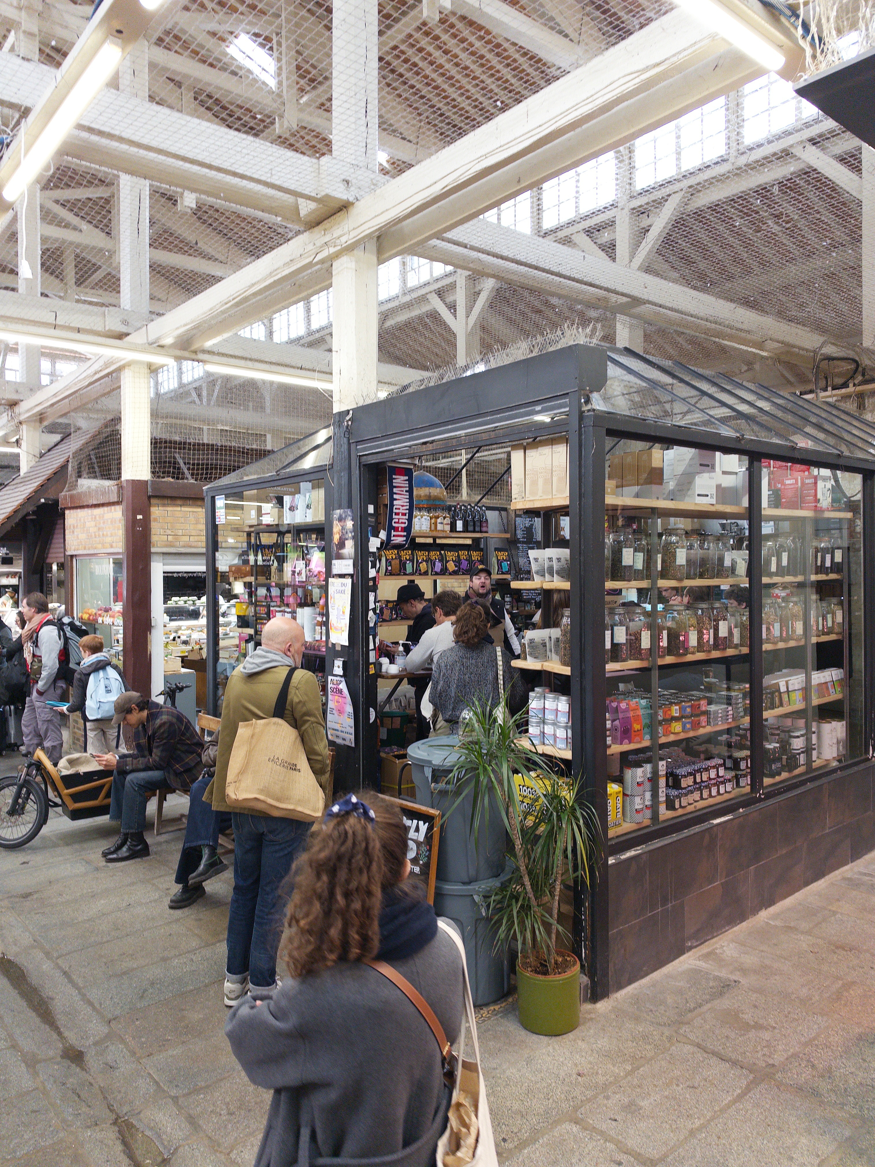 A bustling indoor market stall showcases a variety of products while people wait in line and browse nearby.
