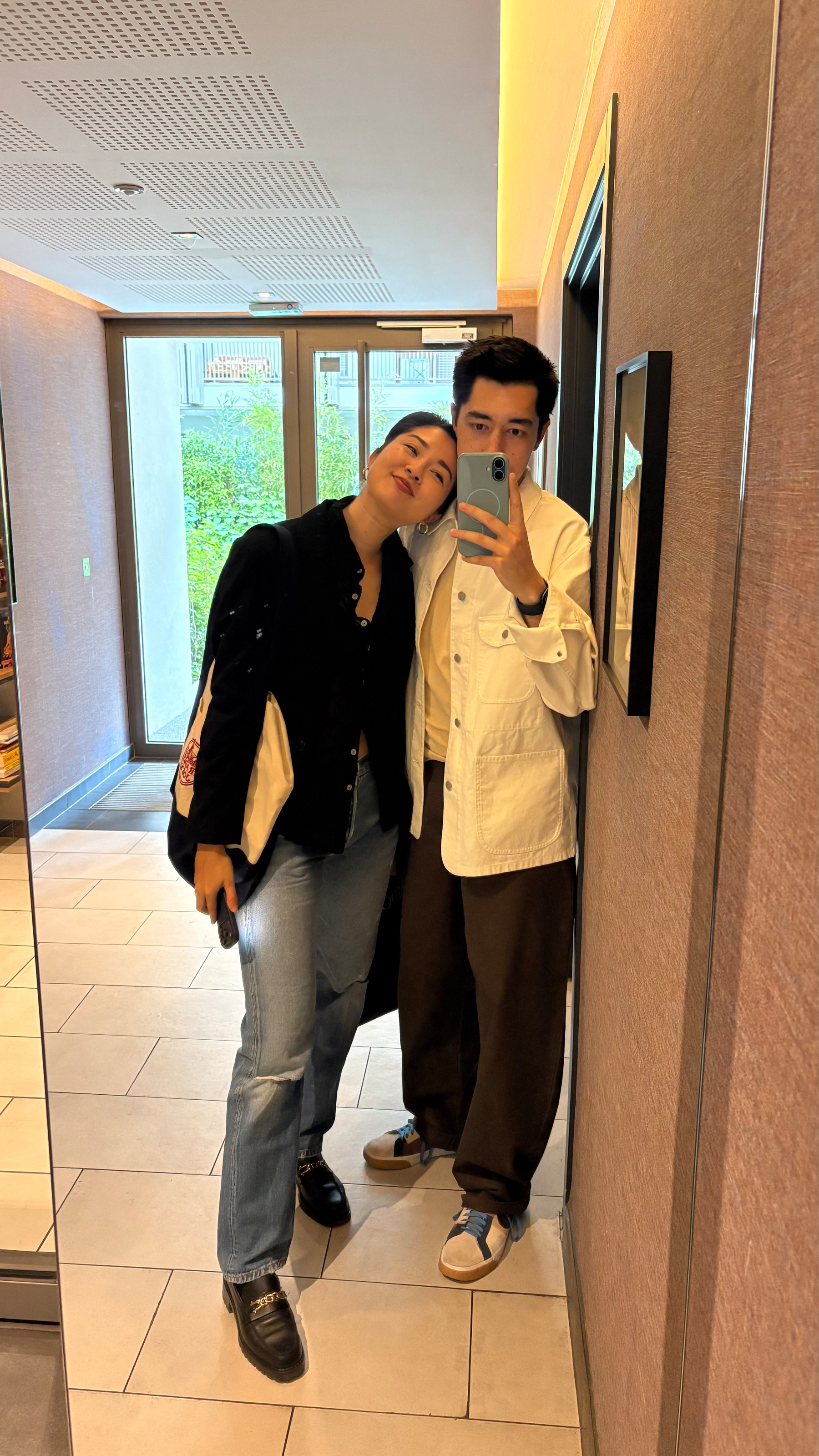 Auto-generated description: Two people are taking a mirror selfie in a hallway, dressed casually, with one person leaning on the other.
