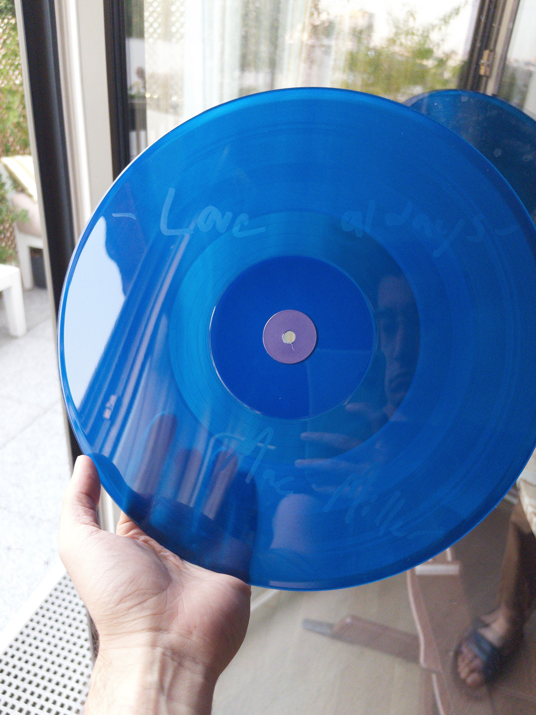 A hand is holding a translucent blue vinyl record with faint handwritten text on it, reflecting the surroundings including a glass door and a person’s face.