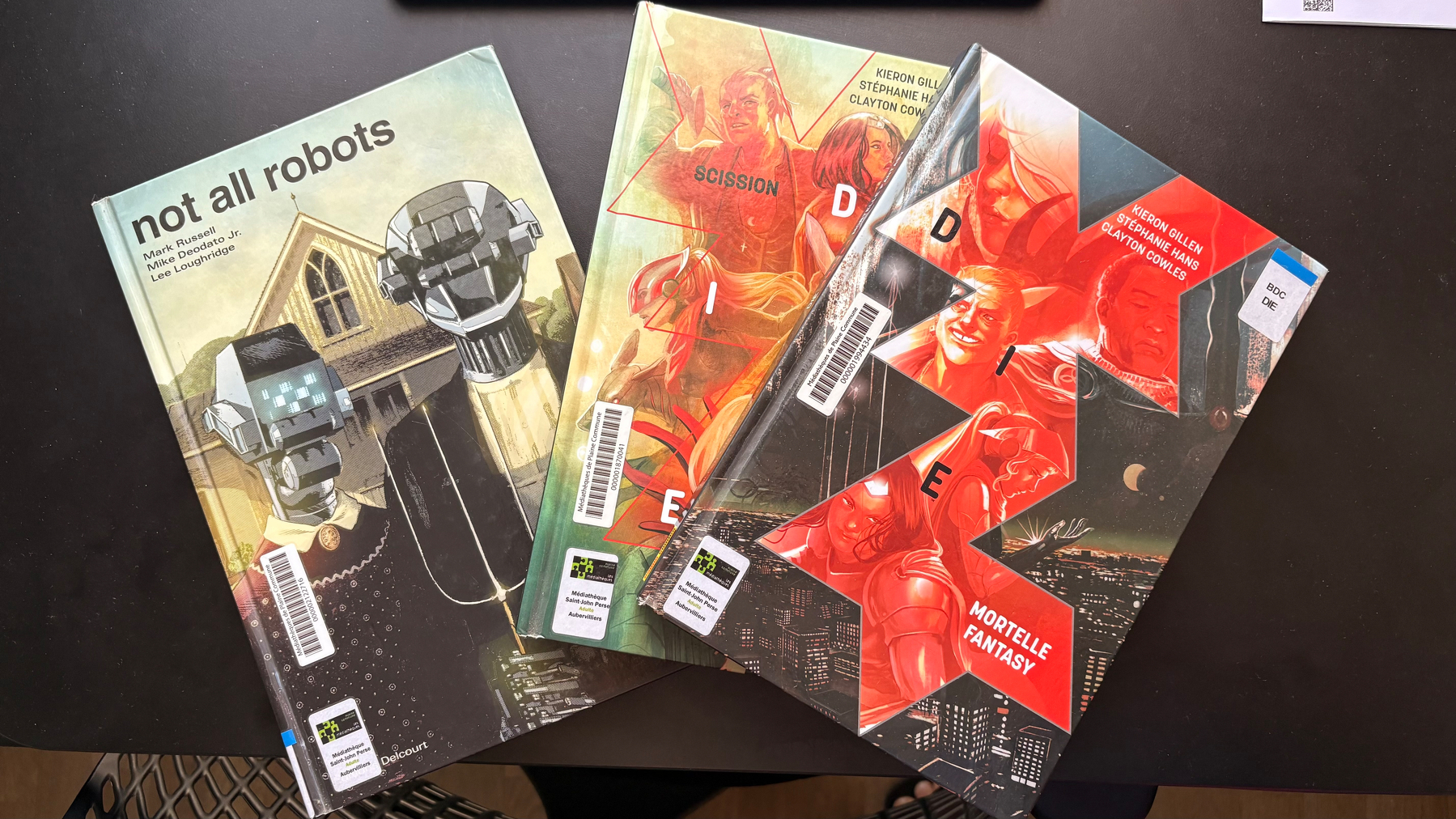 A collection of books is spread out on a dark surface, featuring titles such as not all robots and DIE with colorful, illustrated covers.