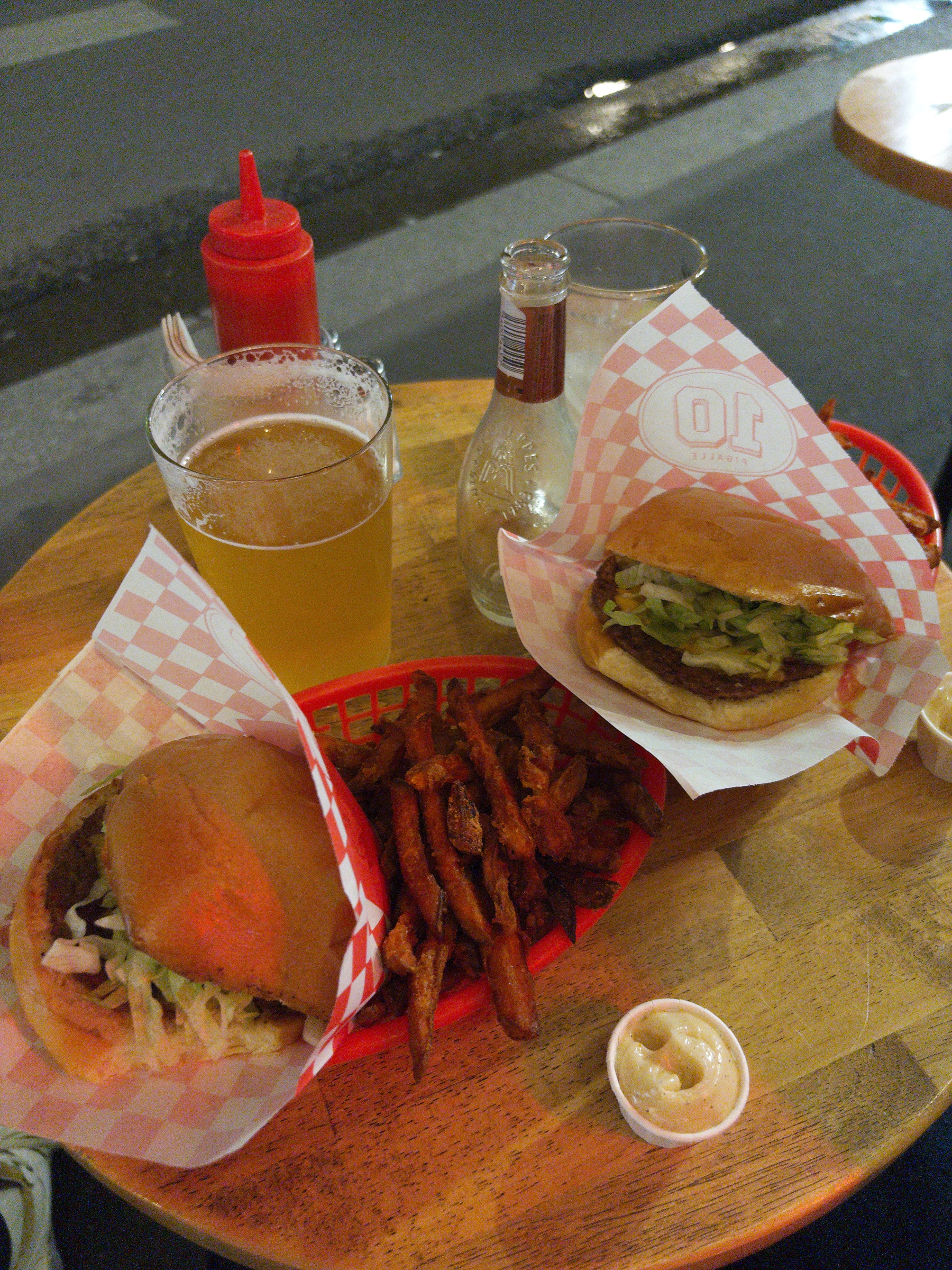 Auto-generated description: A wooden table is set with two burgers in red baskets, a serving of fries, a glass of beer, a soda bottle, ketchup, and a condiment cup.