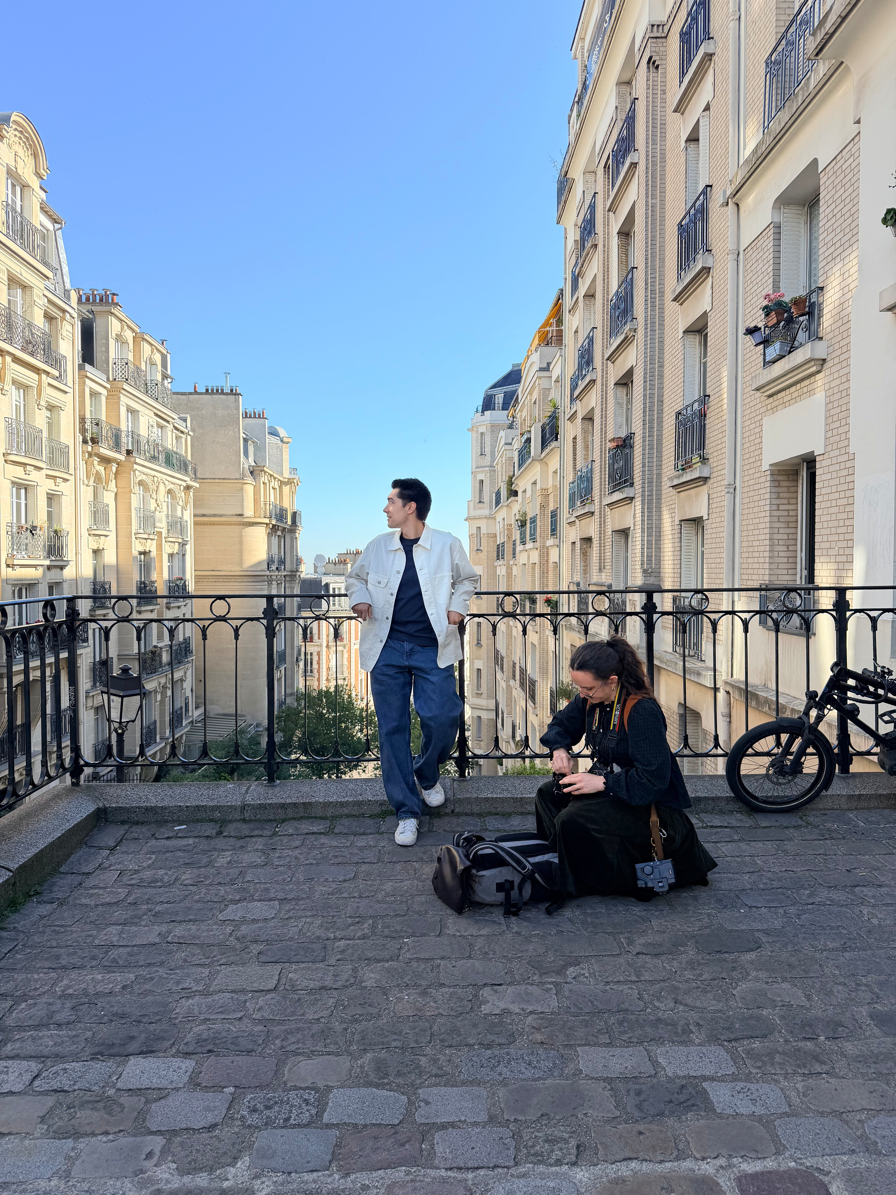 Auto-generated description: Two people relax on a cobblestone terrace, with bicycles nearby and classic European buildings in the background.
