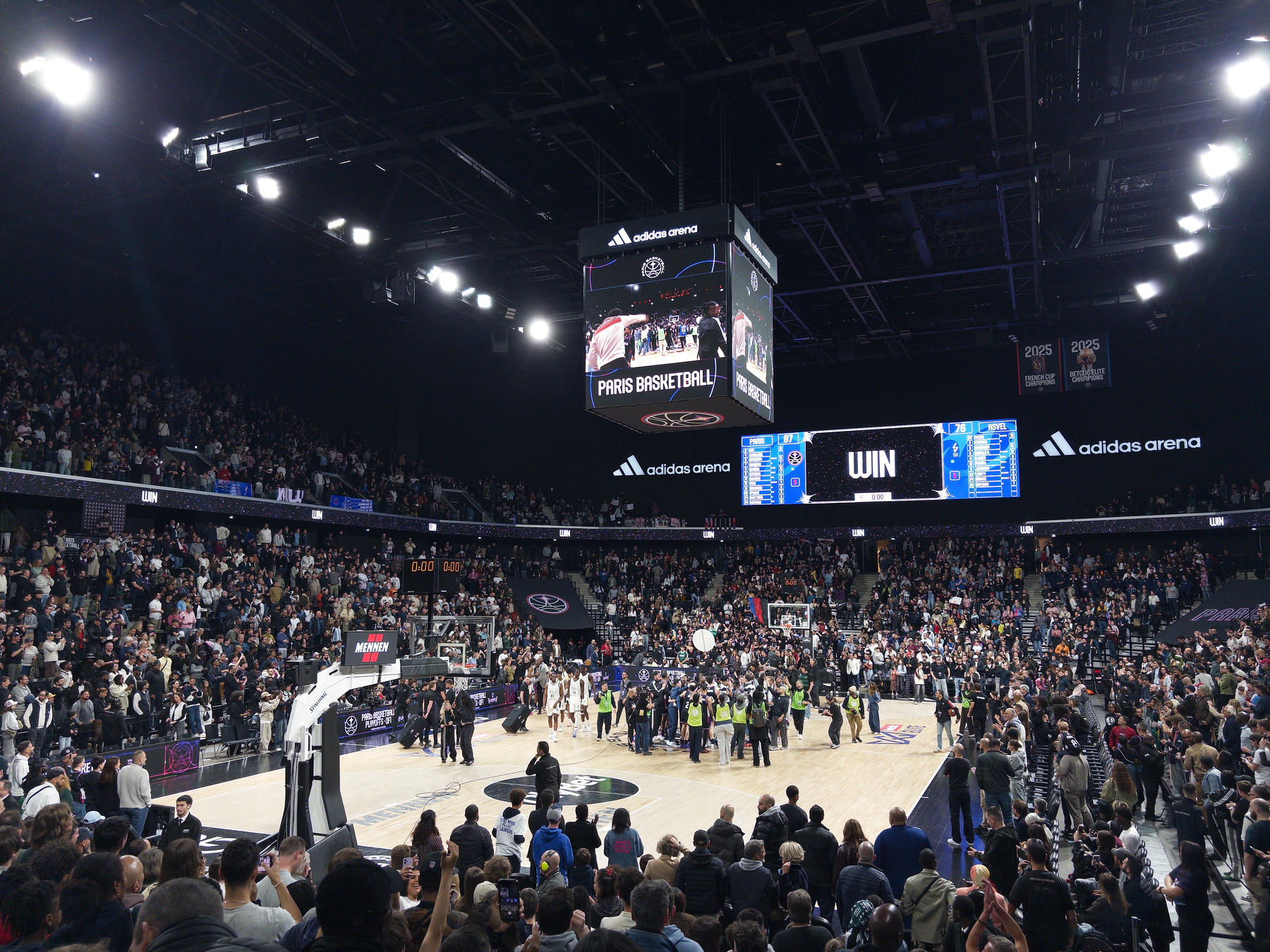 A crowd-filled indoor basketball arena features a large scoreboard and people gathered on the court, celebrating.
