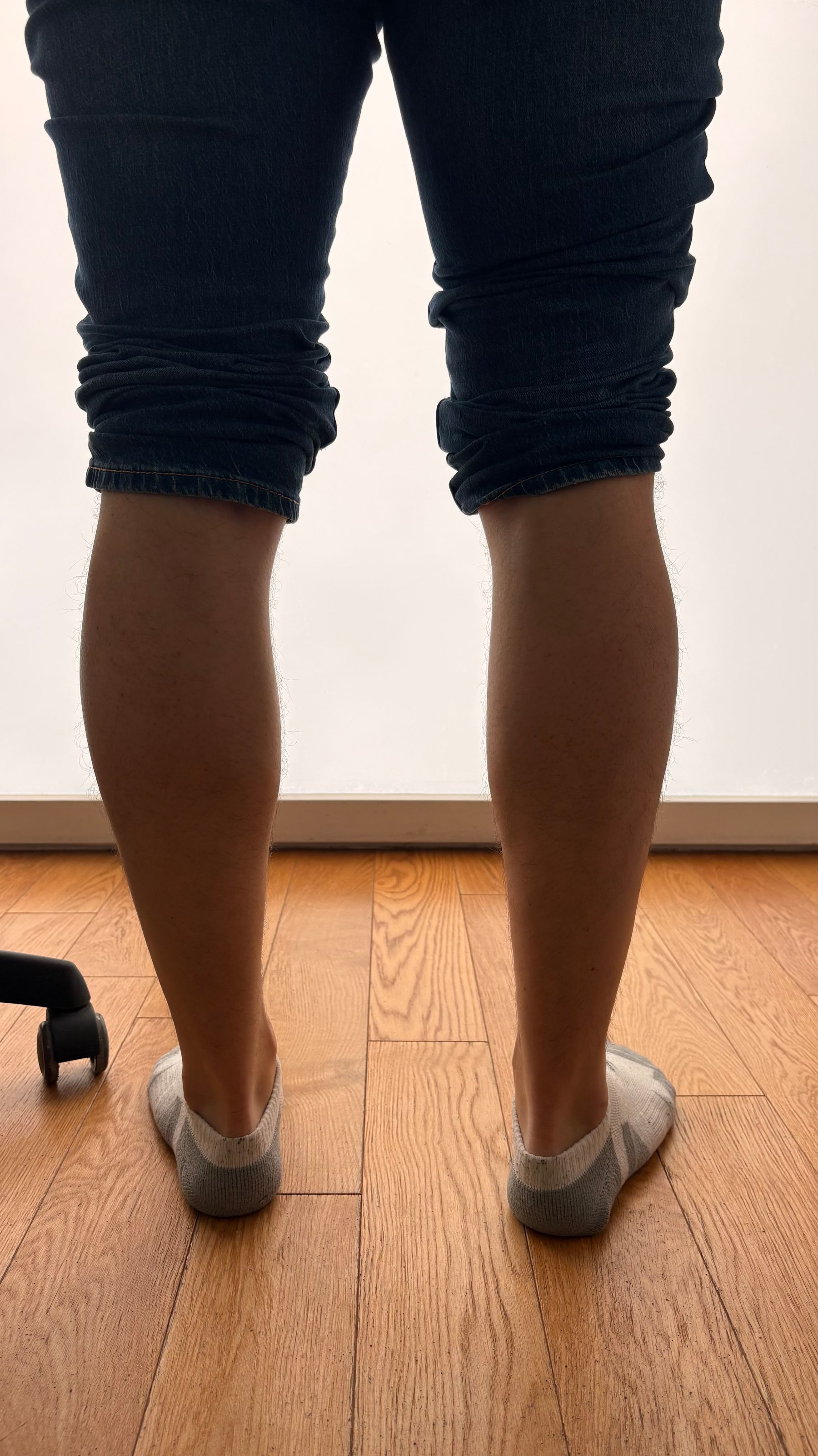 A person wearing rolled-up jeans and gray socks stands on a wooden floor next to a chair wheel.