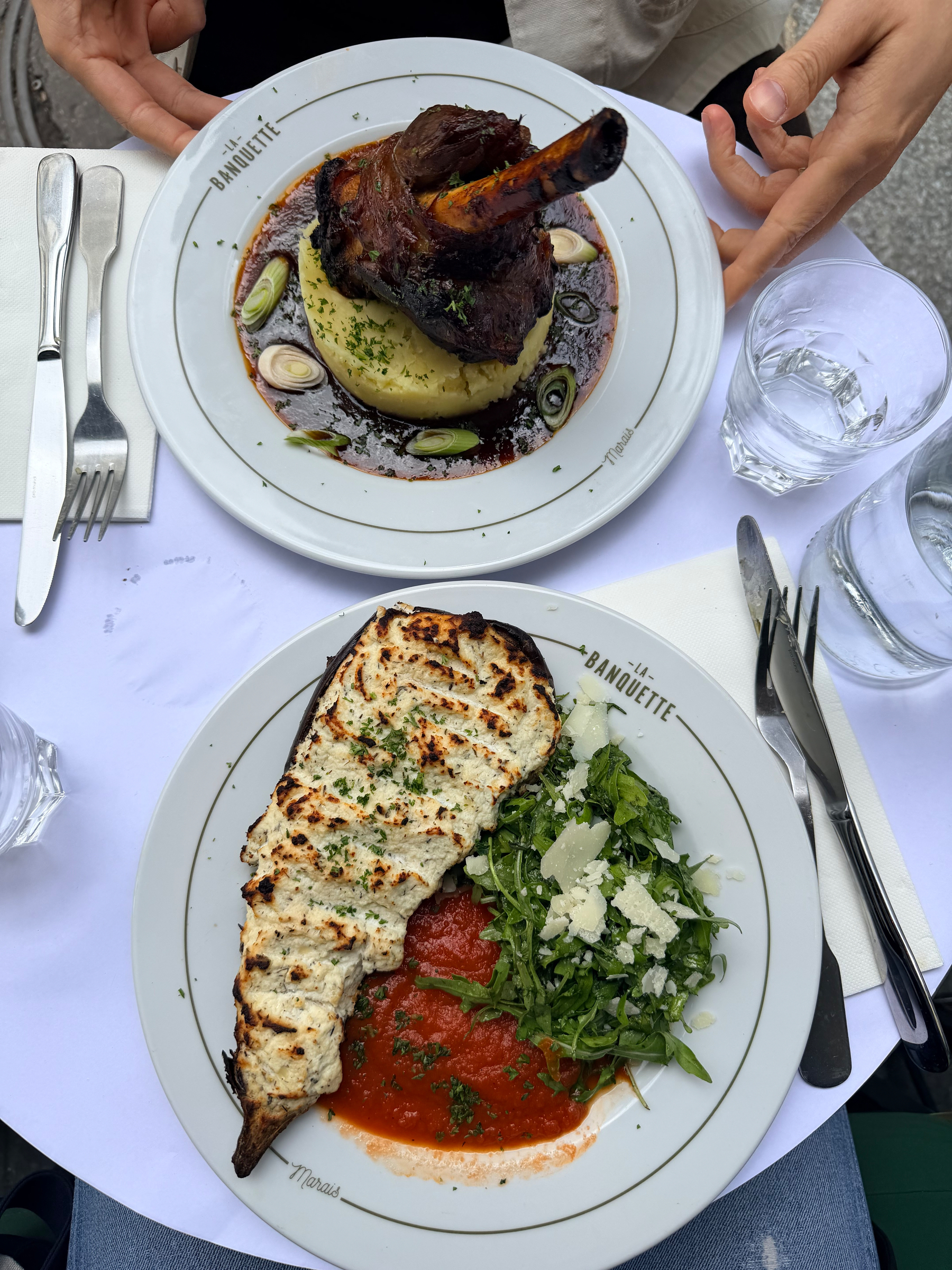 Auto-generated description: Two plates of food are on a table: one with a bone-in meat dish atop mashed potatoes with sauce, and another with a stuffed eggplant alongside greens and tomato sauce.