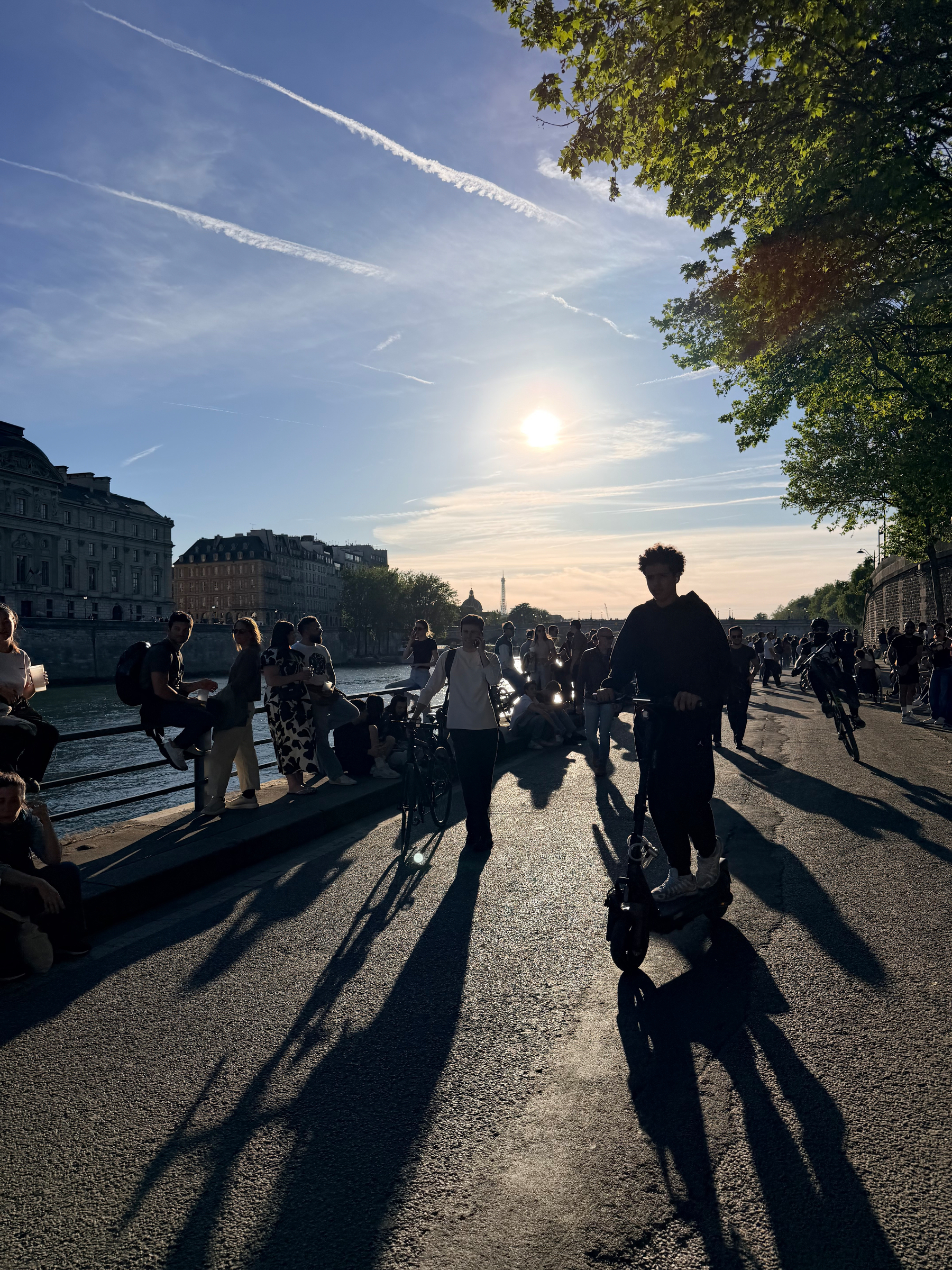People are enjoying a sunny day along a riverside promenade, with some riding scooters and others sitting by the river.