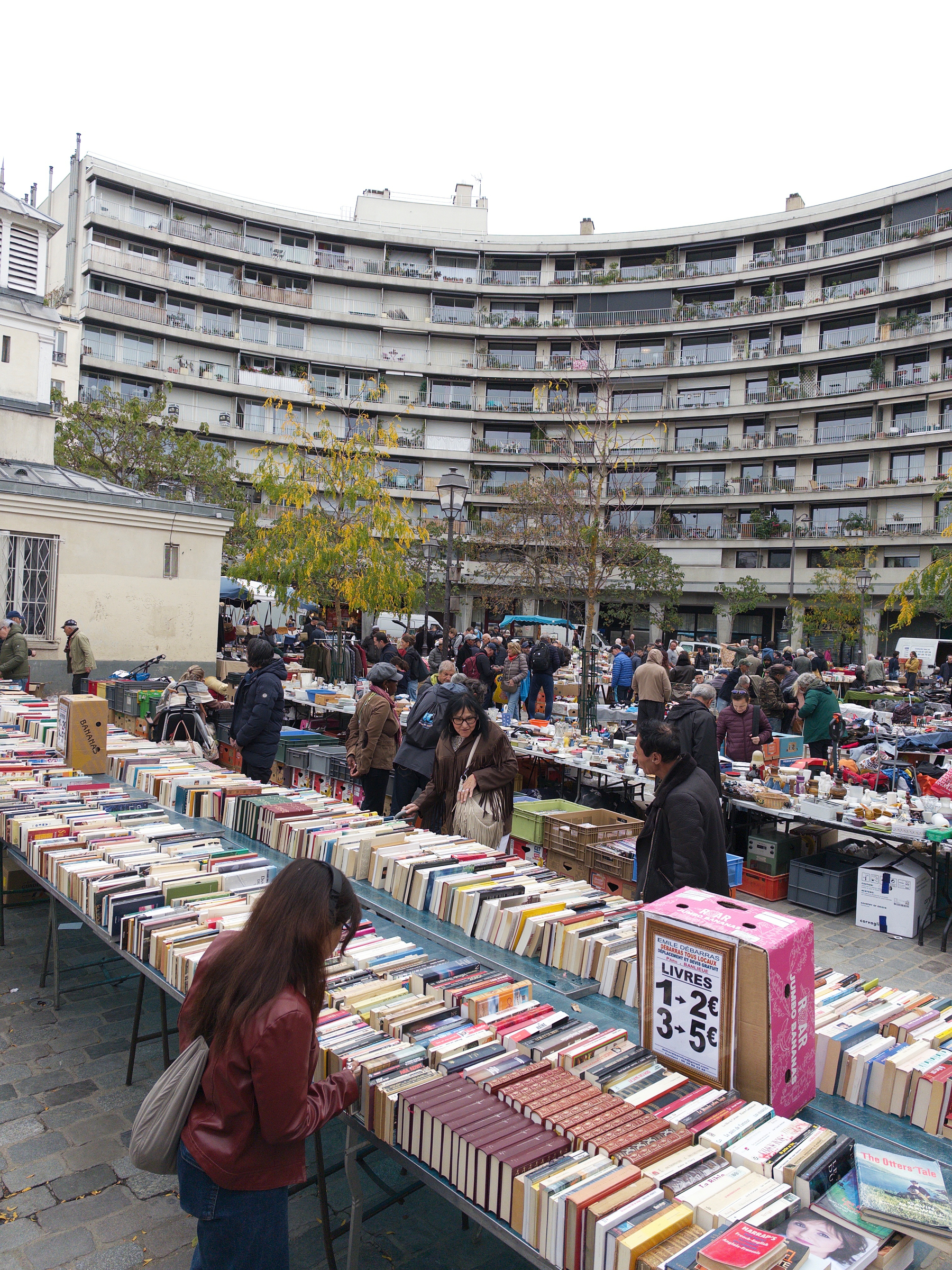 A bustling outdoor book market is set up in a courtyard surrounded by a large curved apartment building, with people browsing through numerous tables of books.