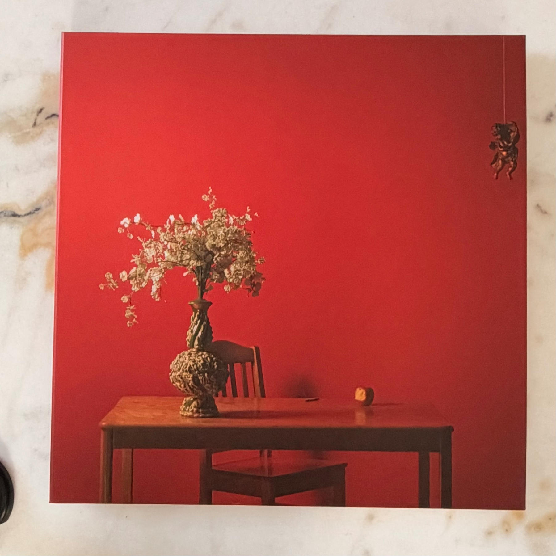 Auto-generated description: A wooden table with a chair behind it features a large floral vase and a small orange object, set against a bold red background with a small hanging cherub figure in the upper right corner.