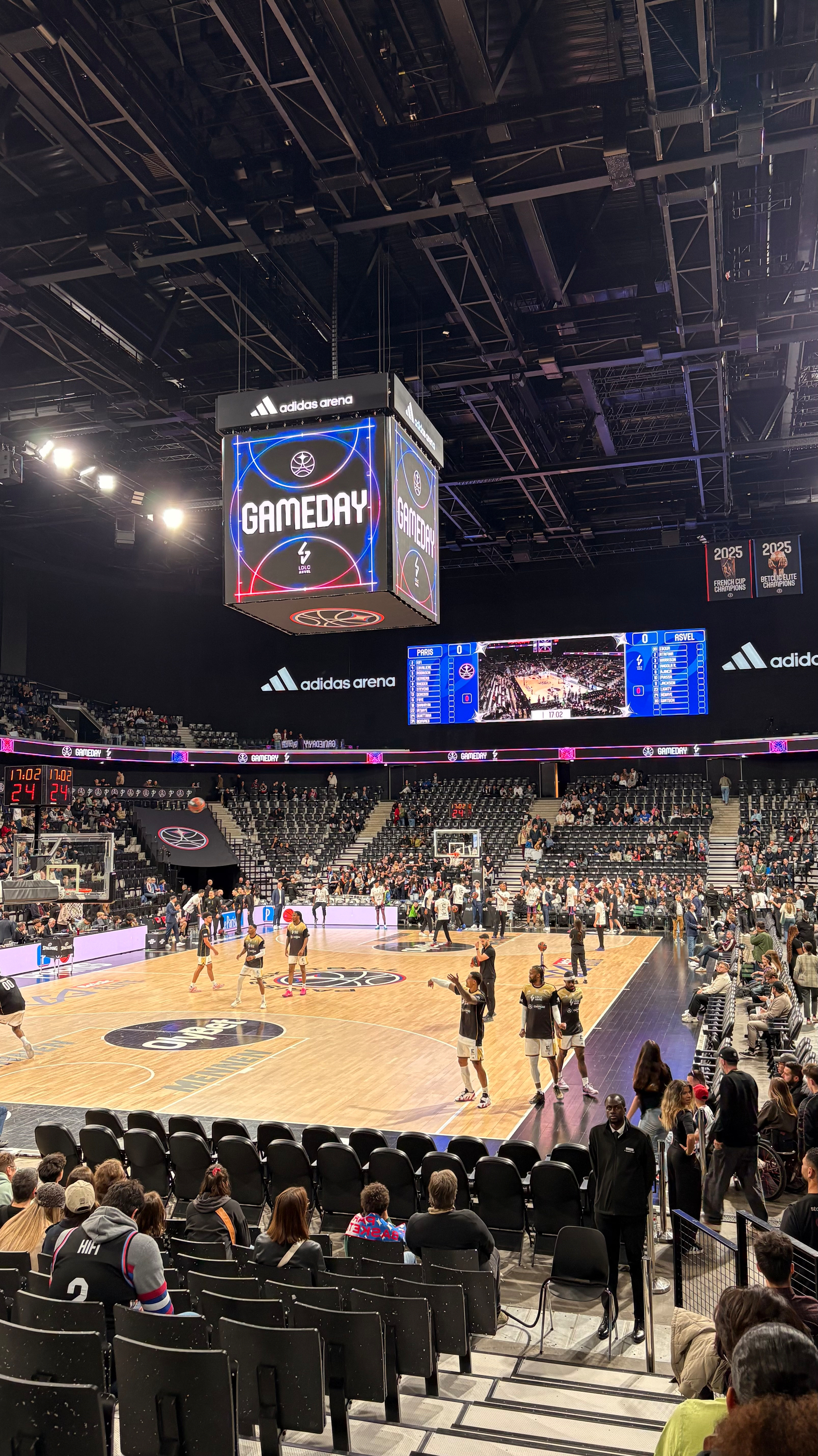 A basketball court inside an arena is bustling with activity, surrounded by spectators and a large overhead scoreboard displaying GAMEDAY.