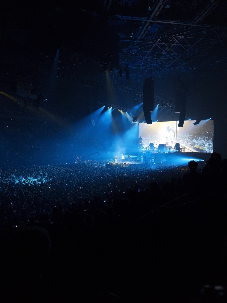A large crowd is watching a live concert with blue stage lighting and a big screen projecting the performance.
