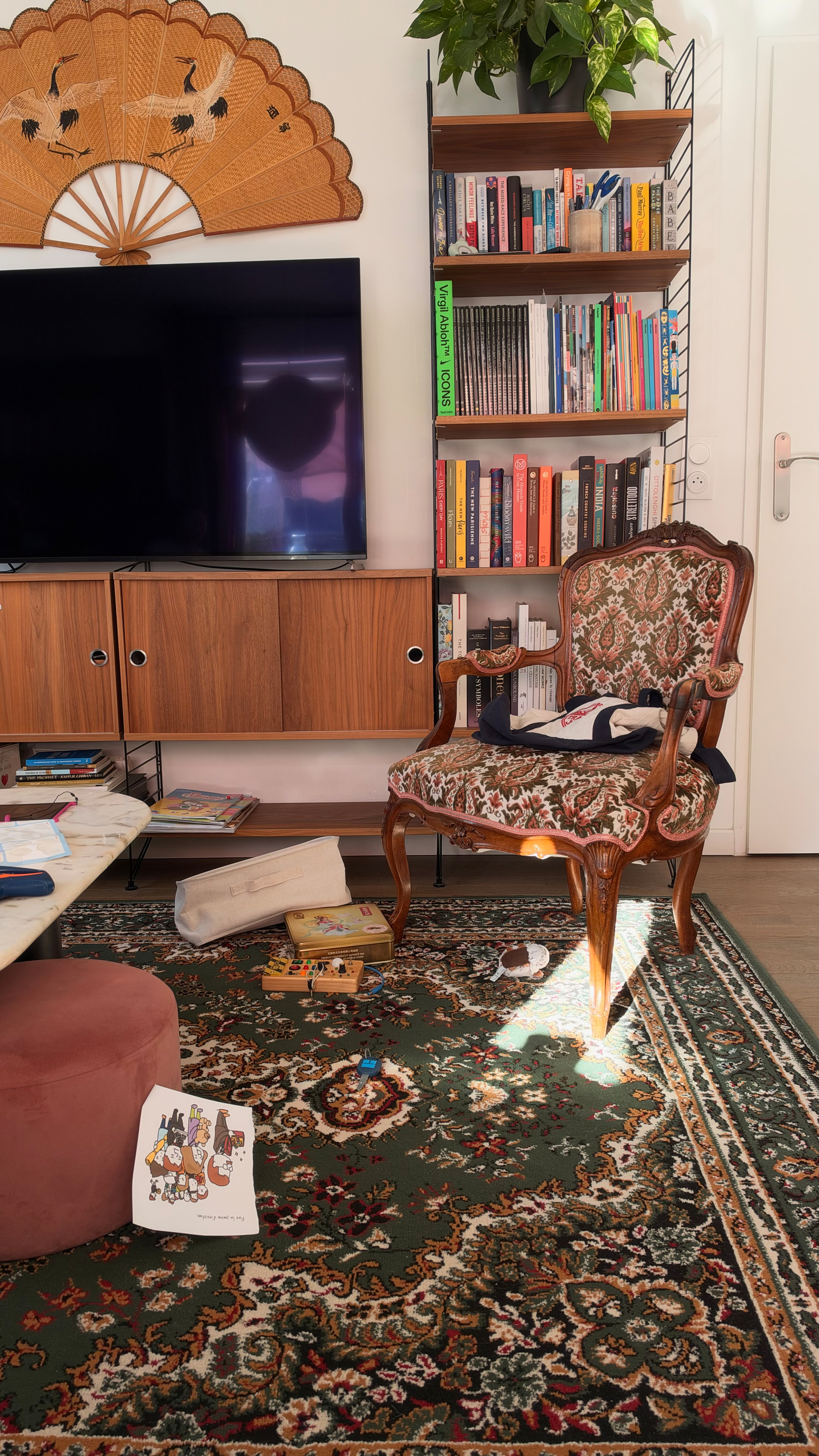 Auto-generated description: A cozy living room corner features a vintage upholstered armchair, a patterned green rug, a wall-mounted TV, wooden shelving filled with books, and a decorative fan with crane designs hanging above the TV.