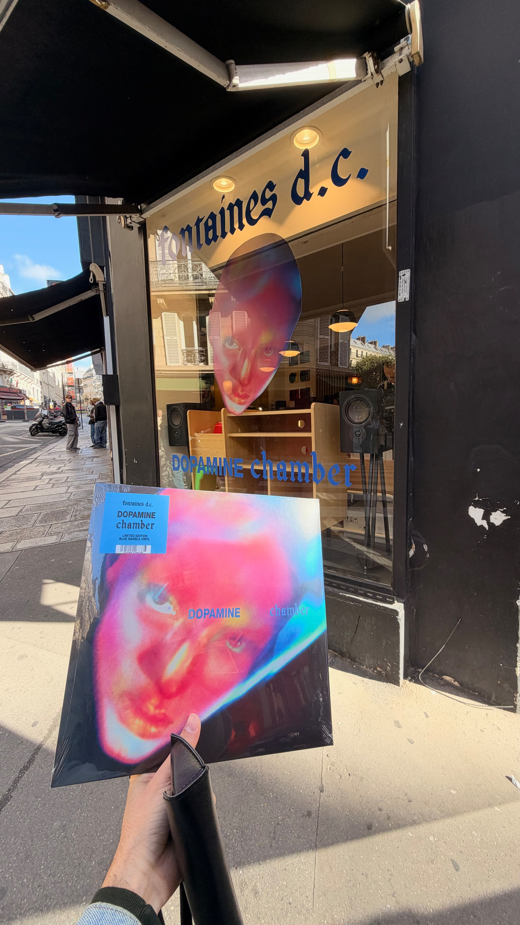 A person is holding a vinyl record with colorful artwork of a face in front of a storefront window displaying the same artwork and text that reads fontaines d.c. DOPAMINE chamber.