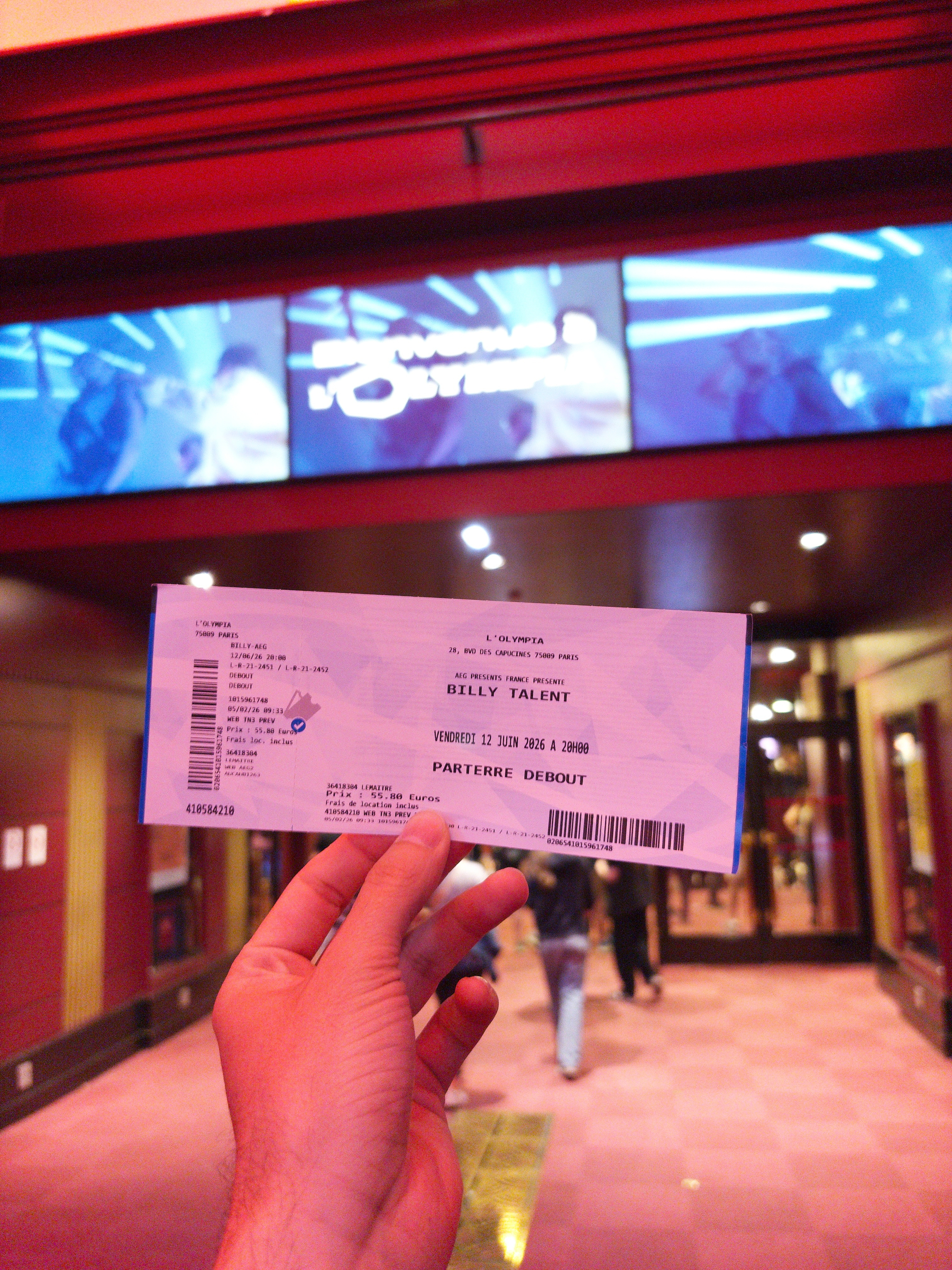 Auto-generated description: A hand is holding a concert ticket for Billy Talent at L'Olympia in front of the venue entrance.