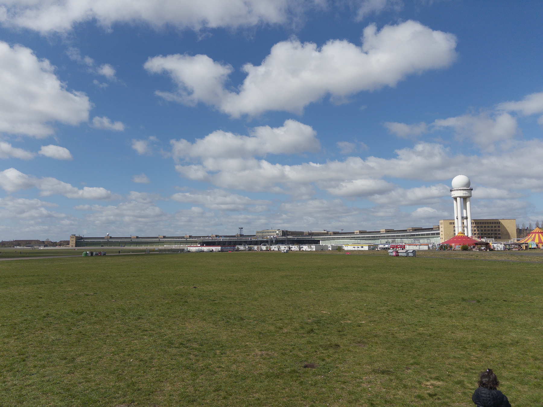 Auto-generated description: A large open grassy field is backed by a building with a white circular structure, under a sky filled with puffy clouds.