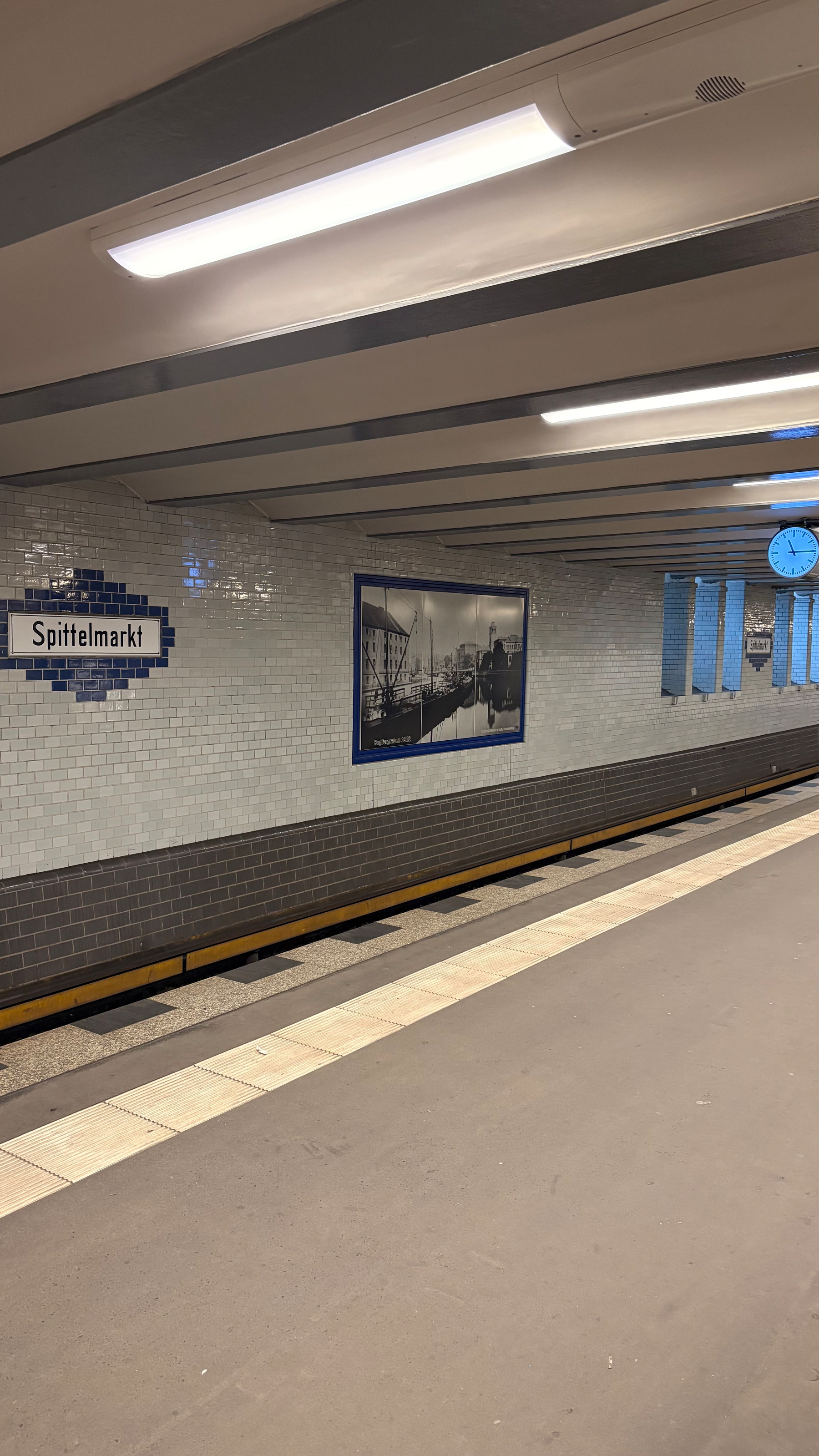Auto-generated description: A subway station platform with a Spittelmarkt sign, a wall clock, and a black-and-white historical photograph.
