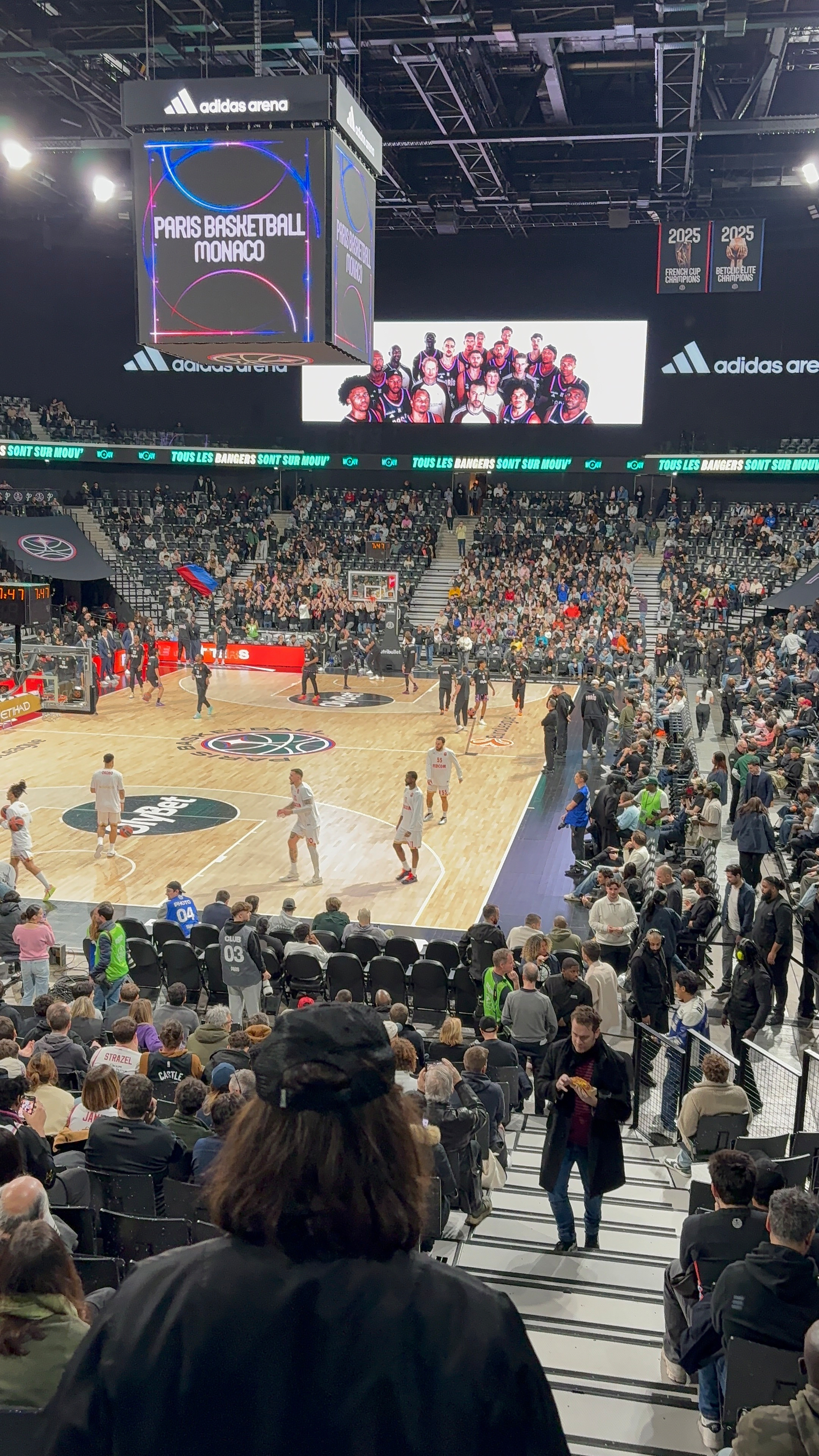 A basketball game is taking place in a large indoor arena filled with spectators, featuring teams warming up on the court.