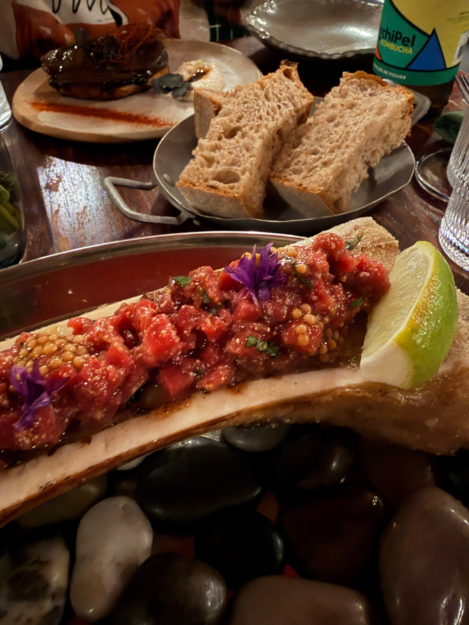 A dish featuring bone marrow topped with a savory minced mixture and garnished with edible flowers is served alongside a lime wedge and accompanied by slices of rustic bread.