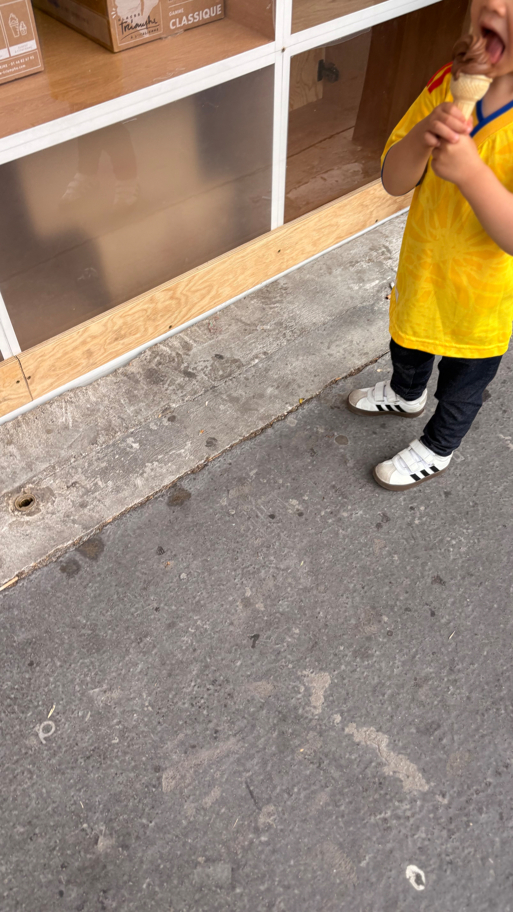 A child wearing a yellow shirt and white shoes is enjoying an ice cream cone on a sidewalk.