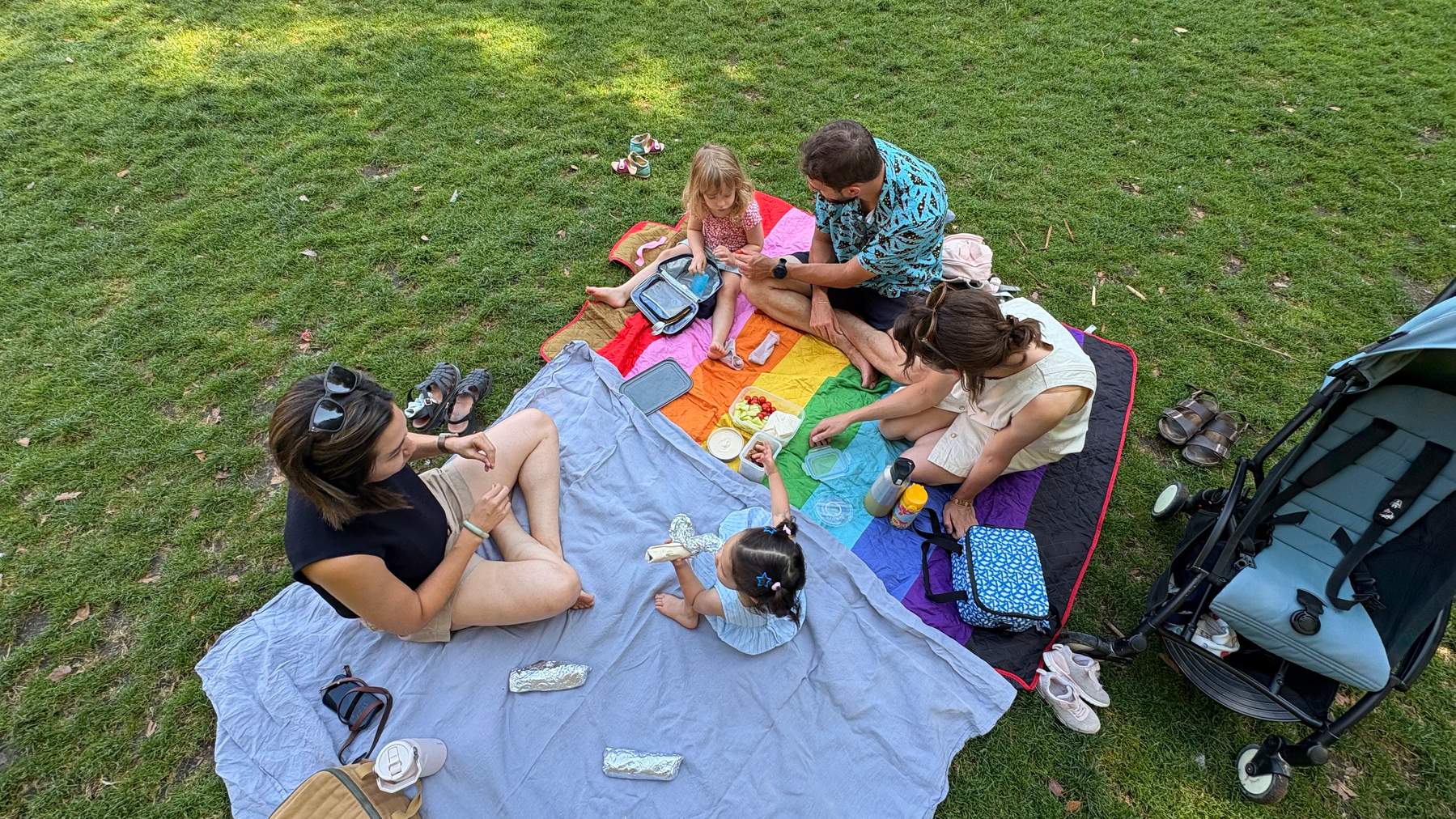 A family is enjoying a picnic on a colorful blanket in a grassy park area with various items and a stroller nearby.