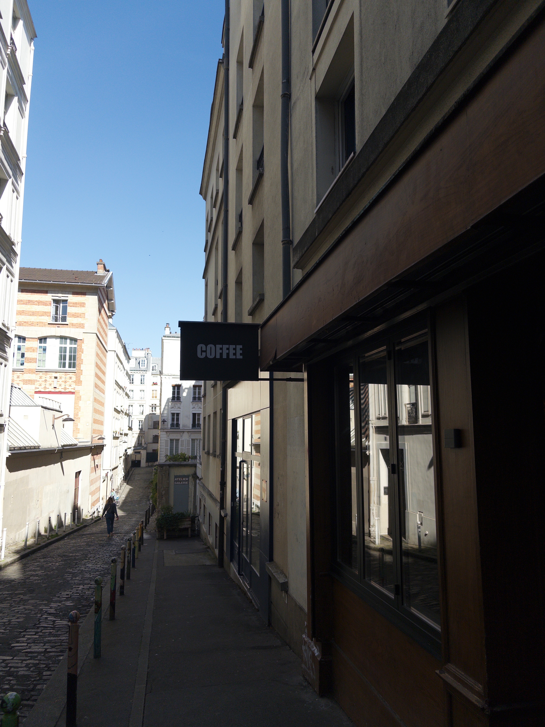 Auto-generated description: A narrow cobblestone street with tall buildings on either side features a sign for a coffee shop.