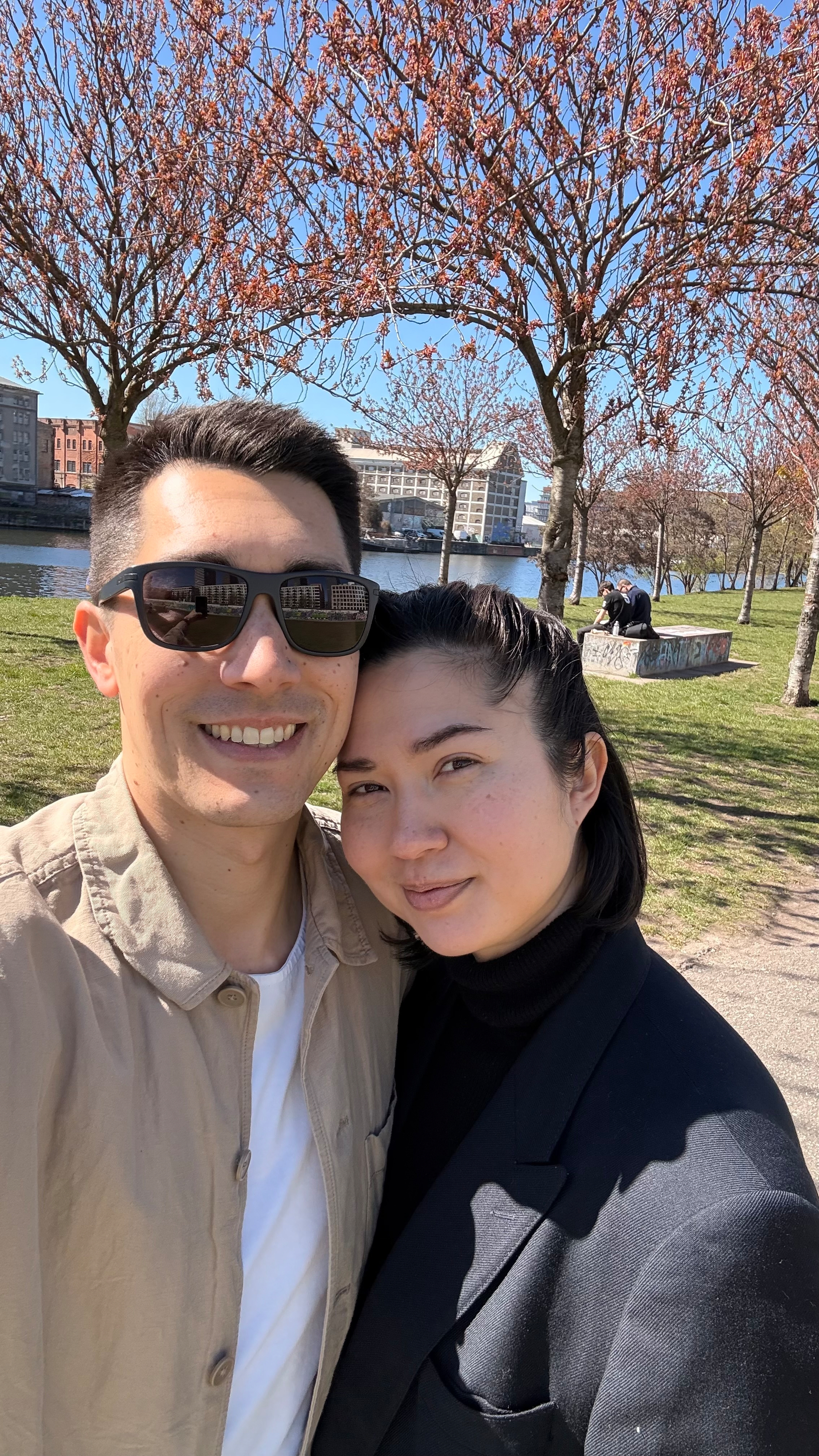 Auto-generated description: A couple poses for a selfie in a park with blossoming trees and a river in the background.